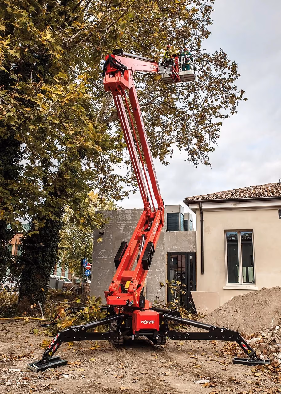 Red spider lift with two workers elevated near tree branches beside construction site.