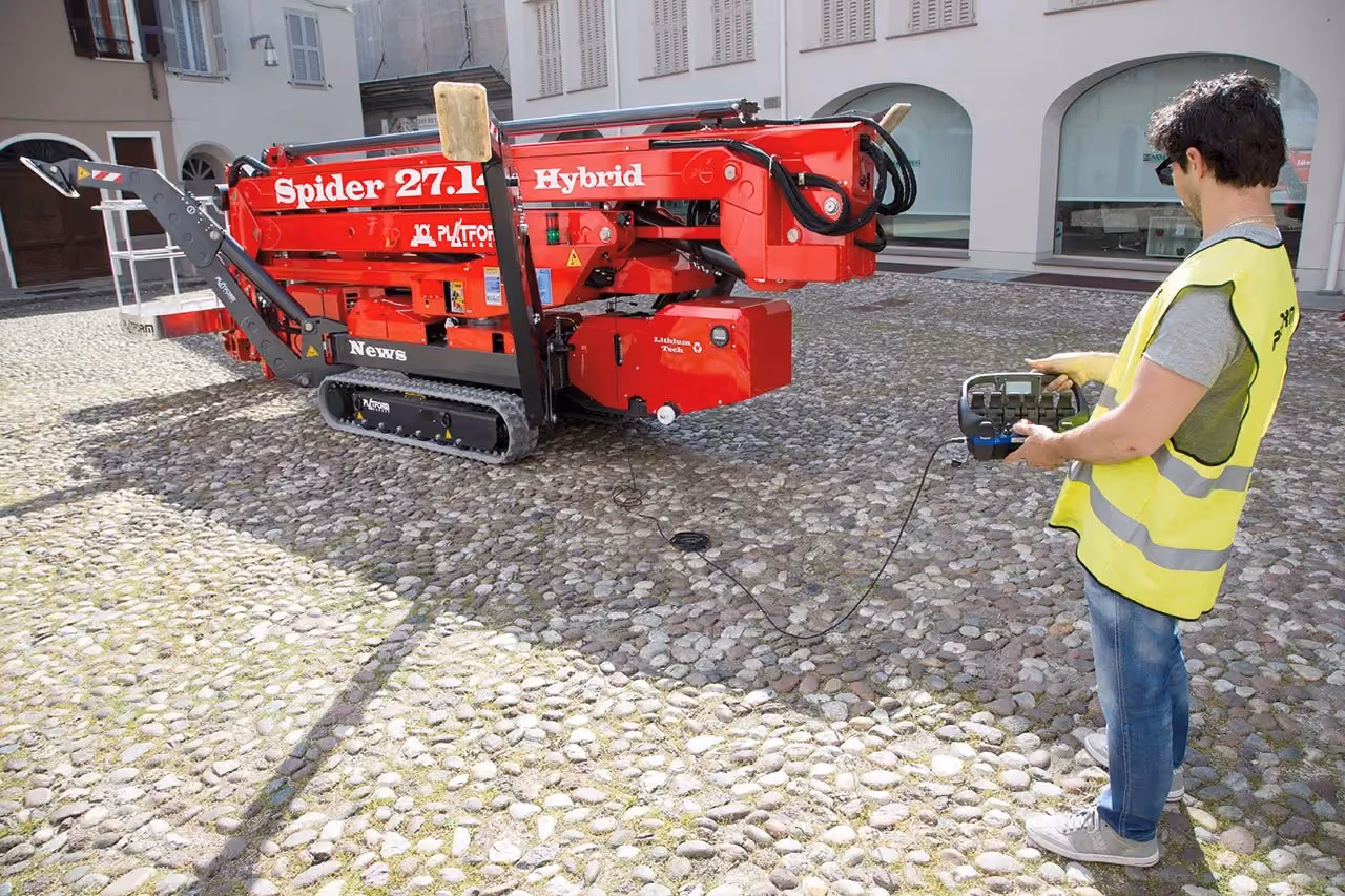 Man in yellow safety vest operates a red Spider 27.1 Hybrid tracked lift on cobblestone courtyard.