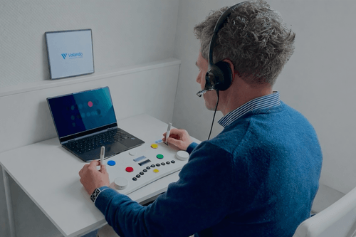 Man wearing headset operating a testing device with colored buttons connected to a laptop at a white desk.