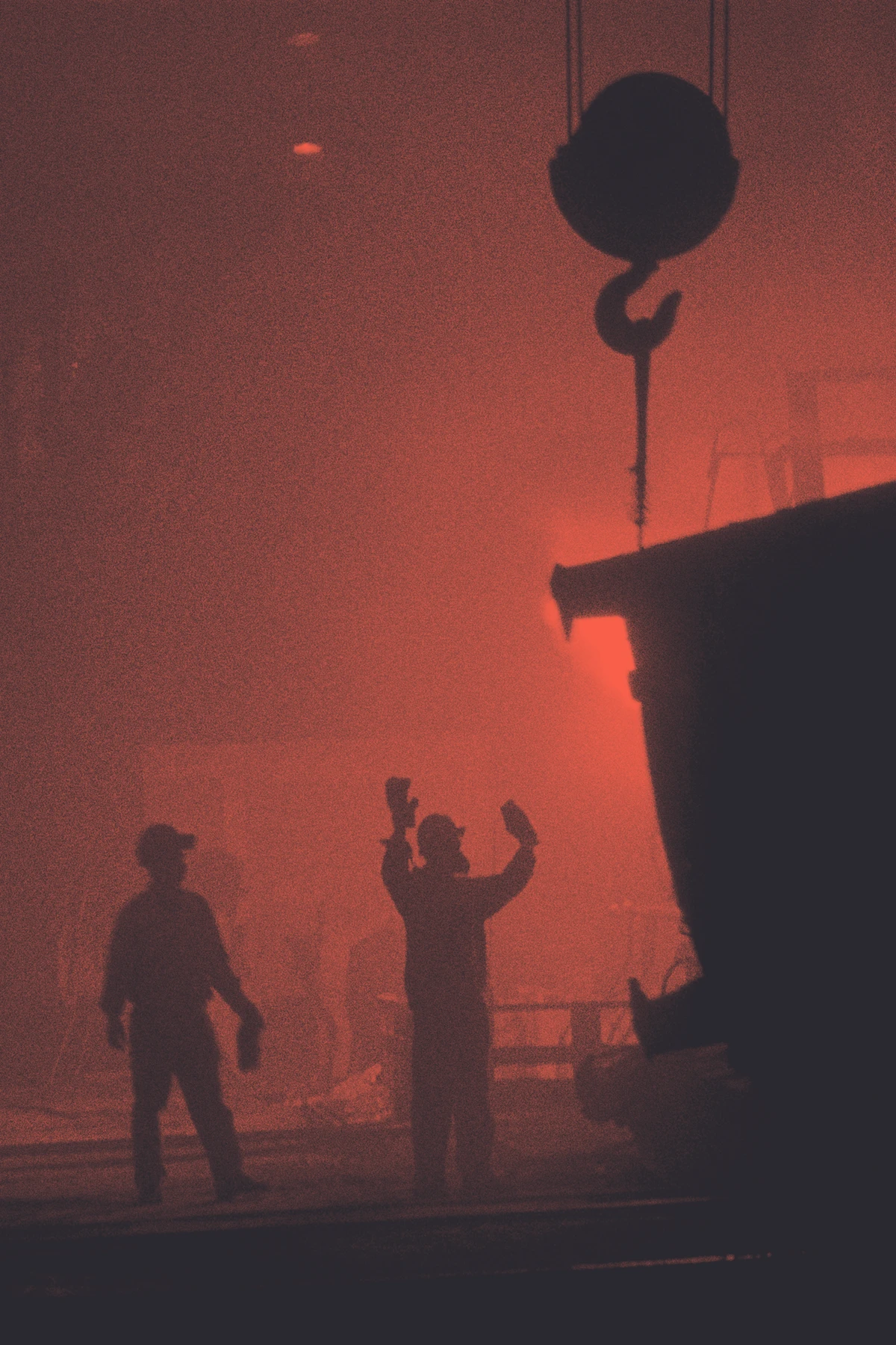 Silhouetted workshop team working in a smoky, industrial space with a large suspended hook overhead.
