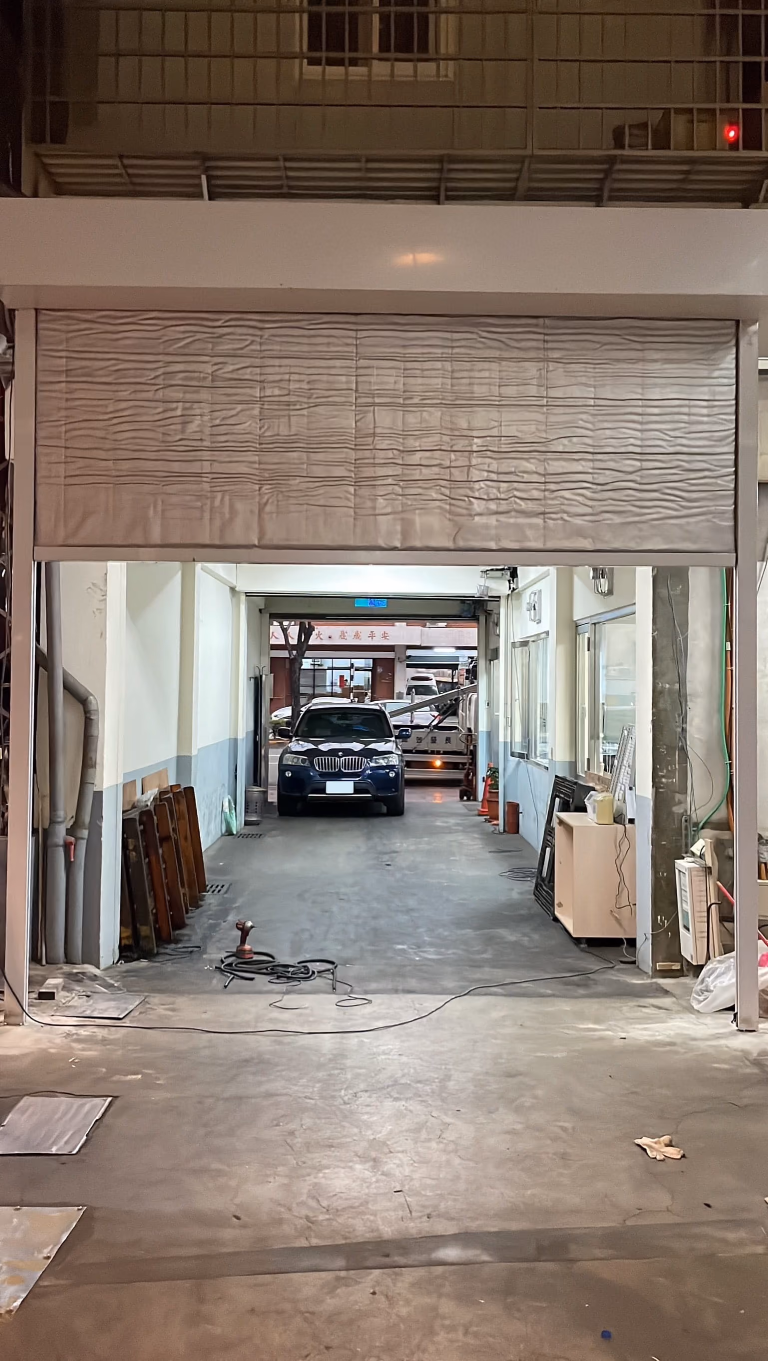 車庫內停放著一輛深色轎車，旁邊有工具和家具，環境為工業或修車空間。