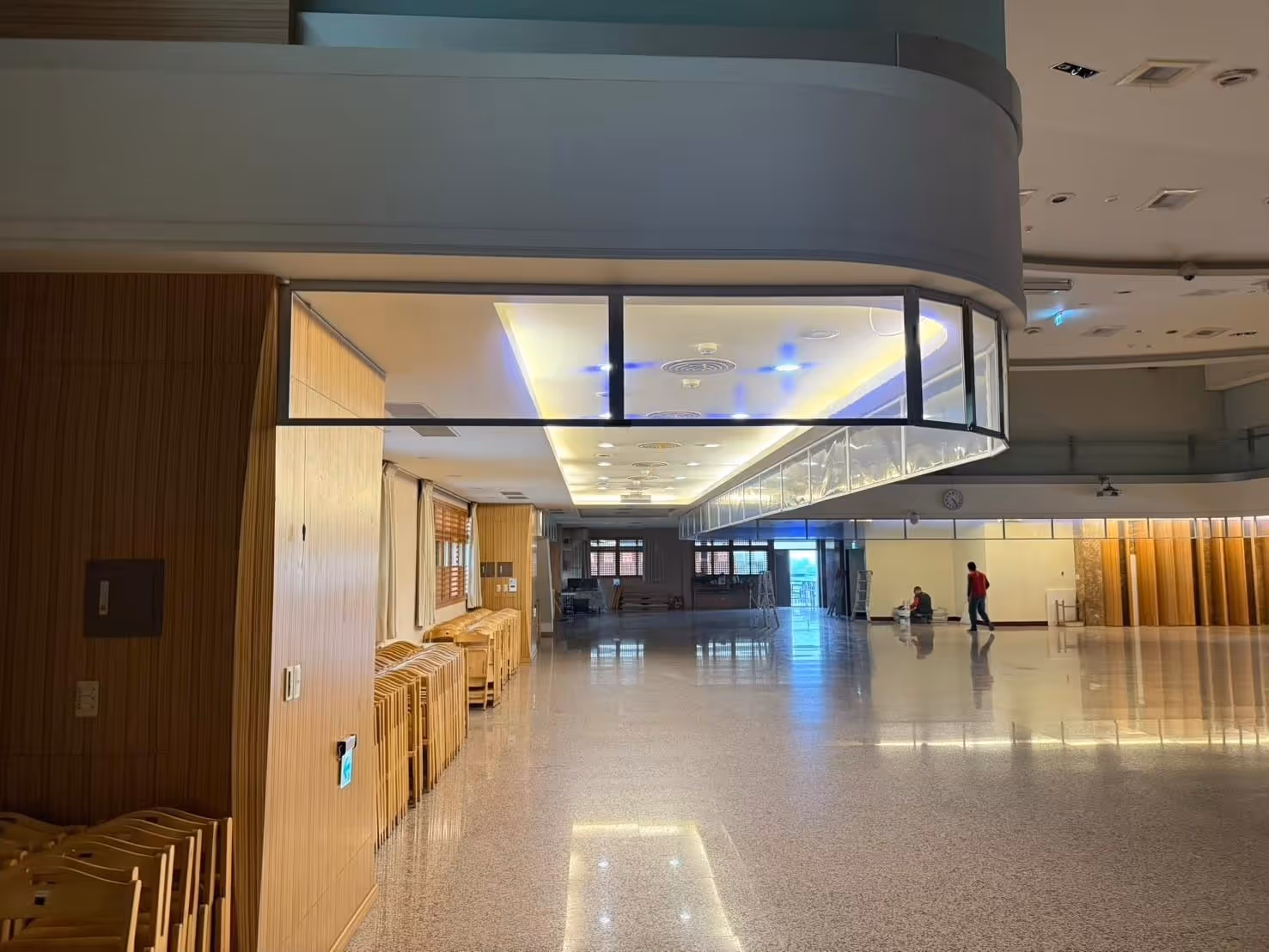 大型空曠室內空間，有堆疊木椅與兩人靠牆休息或走動。