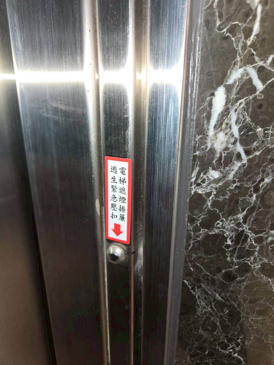 貼在金屬扶手上的標籤，寫著電梯逃生緊急壓扣及向下箭頭。