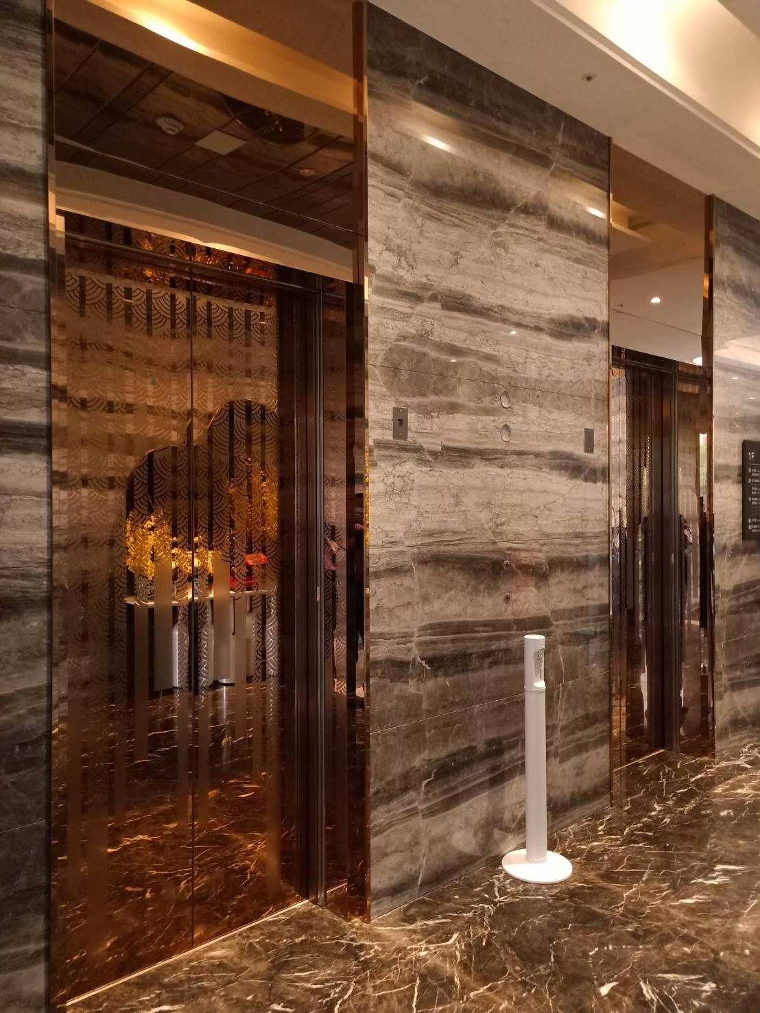 Two elevators with decorative glass doors in a marble-walled lobby with shiny floor and a sanitizer stand.