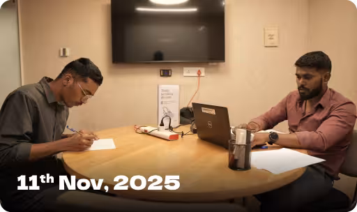 Two men sitting at a round table in a meeting room, one writing on paper and the other using a laptop, with a TV screen mounted on the wall behind them and the date '11th Nov, 2025' displayed.