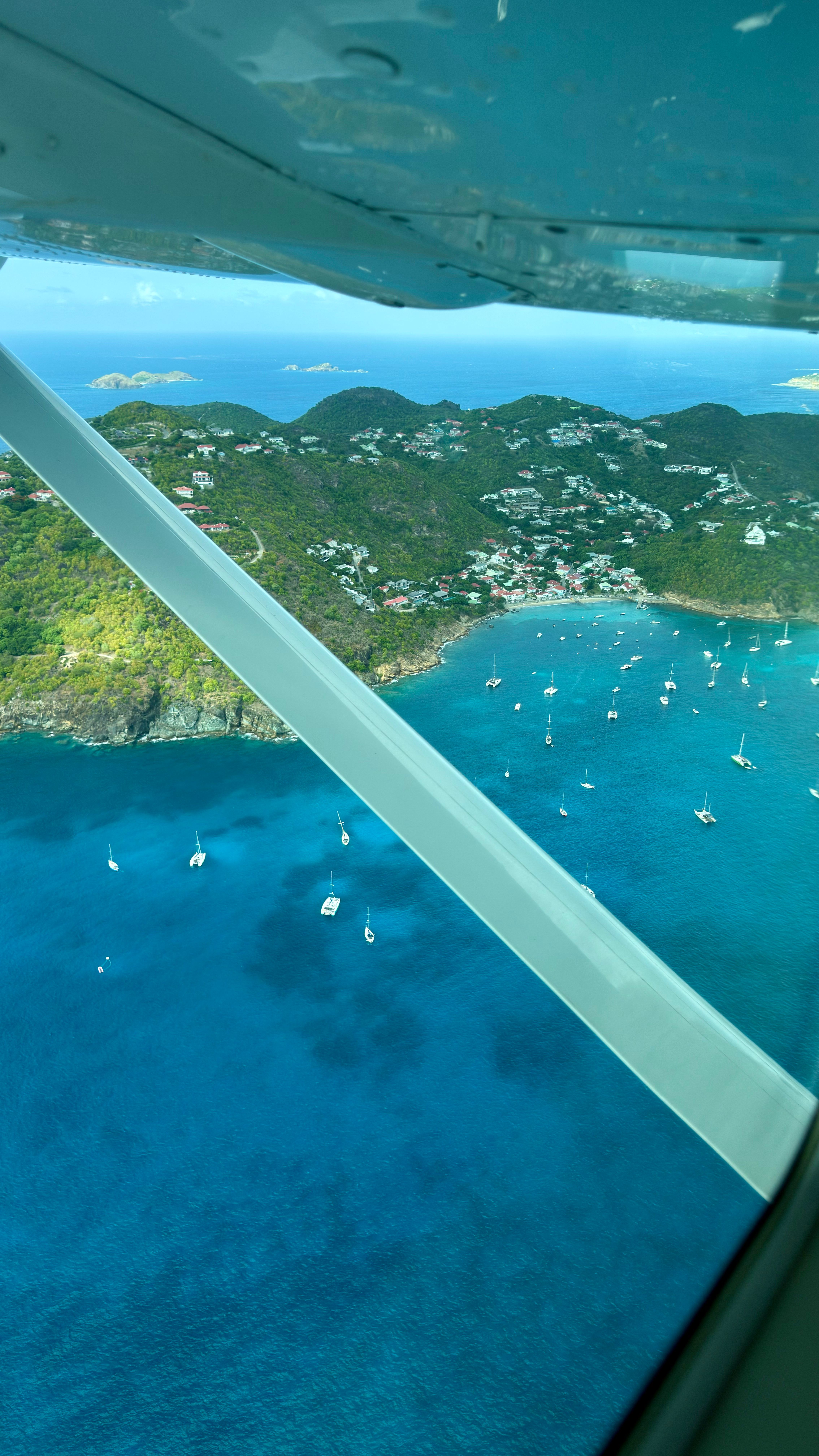 Aerial view from a commuter flight arriving over Saint-Jean Bay in St. Barths with turquoise water and sailboats below.