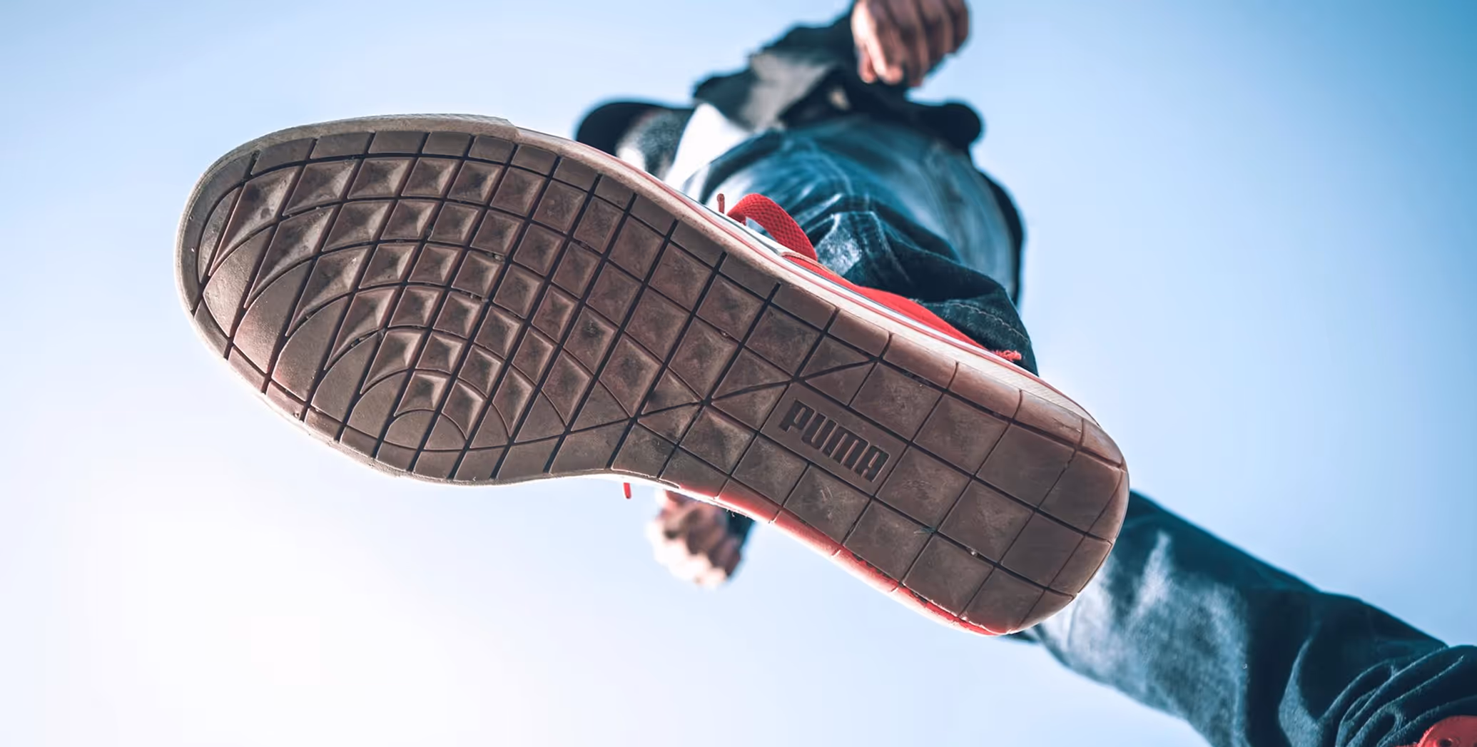 Close-up of the sole of a red Puma sneaker worn by a person walking, with blue sky in the background.