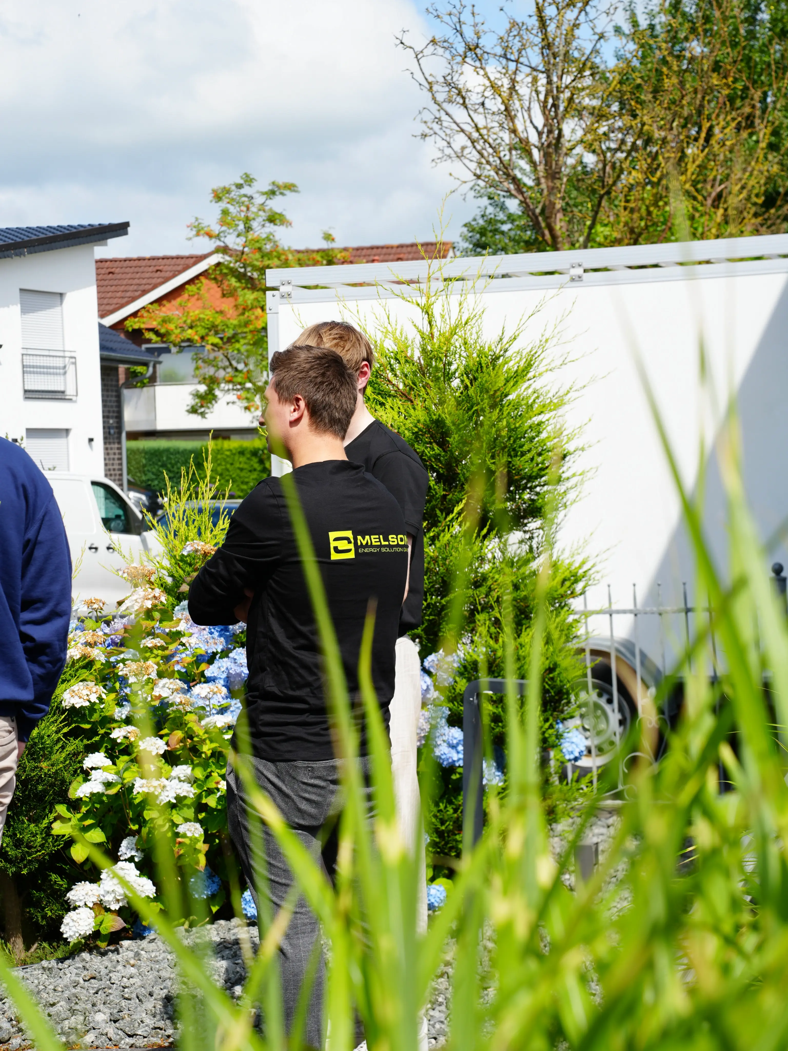 Melson Home Teammitglieder im Gespräch im Vorgarten eines Wohnhauses, mit blühenden Hortensien und Montagefahrzeug im Hintergrund.