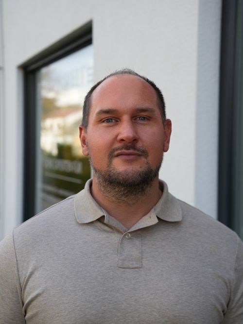 Portrait of a man with short hair and beard wearing a beige polo shirt standing outdoors near a white building.
