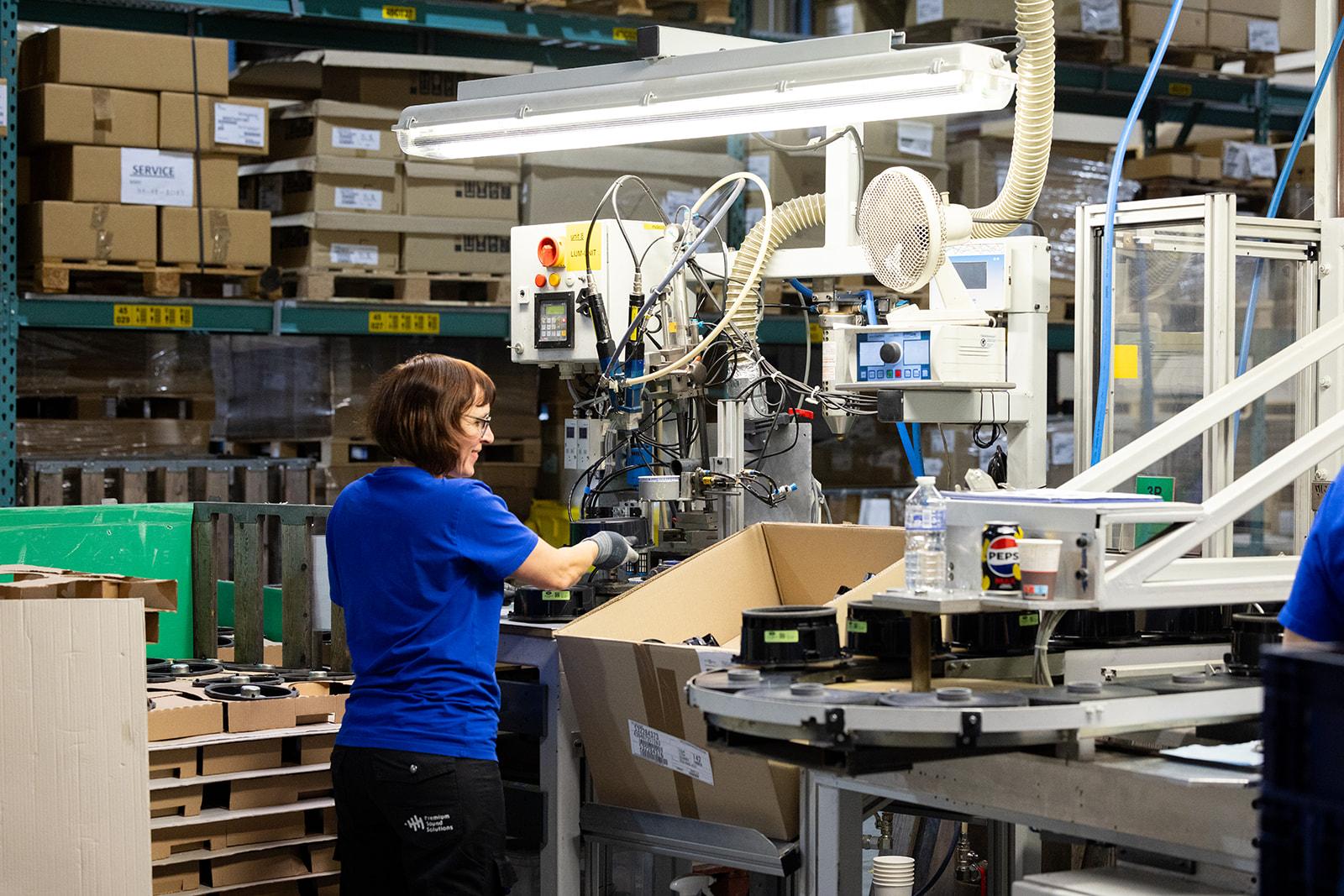 Female factory worker in a blue shirt operating industrial machinery in a warehouse with conveyor belts, cardboard boxes, and various technical equipment.