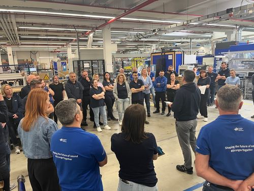 Worker in a high-visibility jacket and gloves secures or adjusts the fastening straps of a large cargo truck, seen through a translucent plastic curtain or reflection.