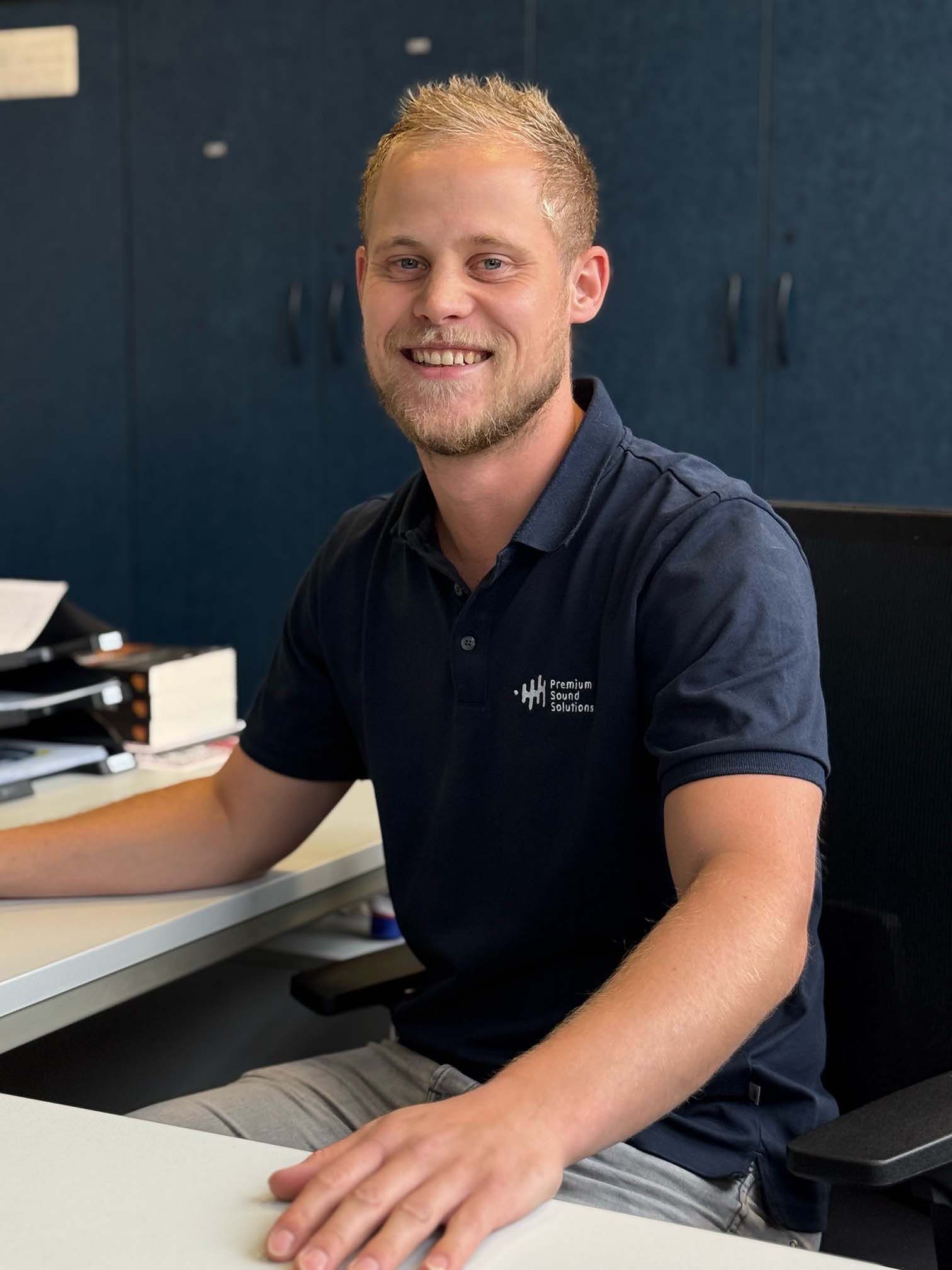 Jonge man met blond haar en baard die glimlacht achter zijn bureau, gekleed in een donkerblauw Premium Sound Solutions polo, kantooromgeving op de achtergrond.