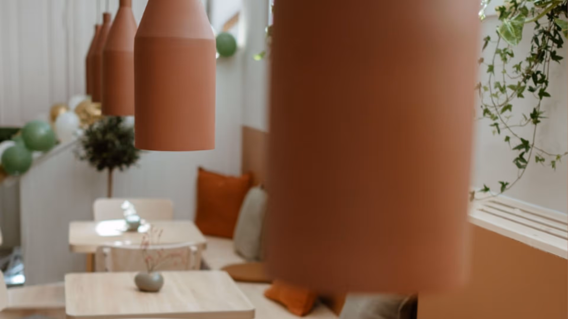Cozy indoor seating area with wooden tables, brown hanging lamps, orange and beige cushions, and green plants.