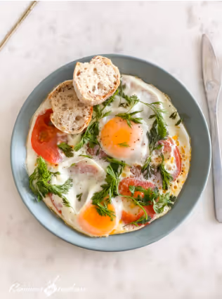 Plate with two fried eggs garnished with fresh dill and sliced tomatoes, served with two pieces of bread on the side and a knife placed on the right.