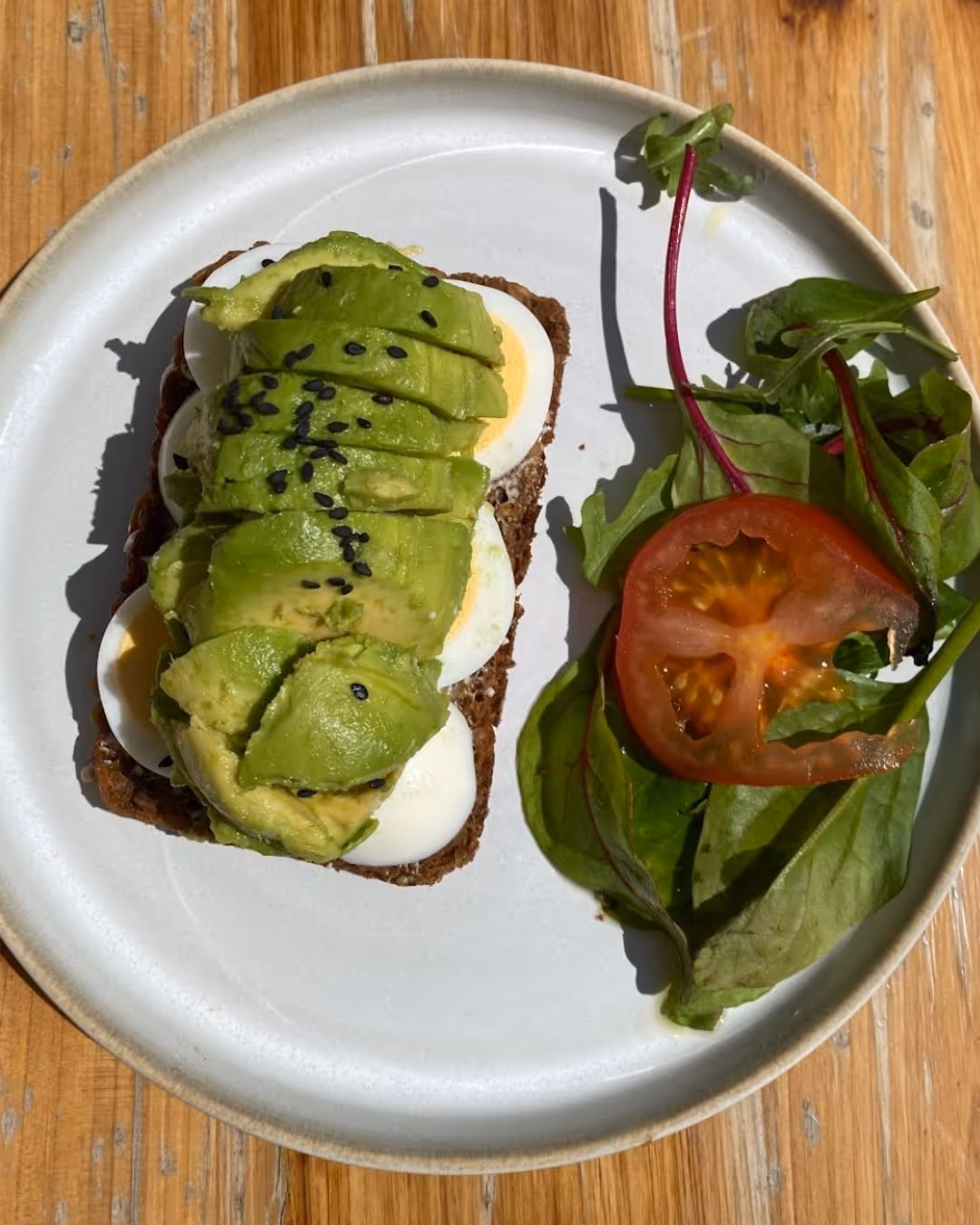 Sandwich with sliced avocado, hard-boiled egg, and black sesame seeds on whole grain bread, served with mixed greens and a tomato slice on a white plate.