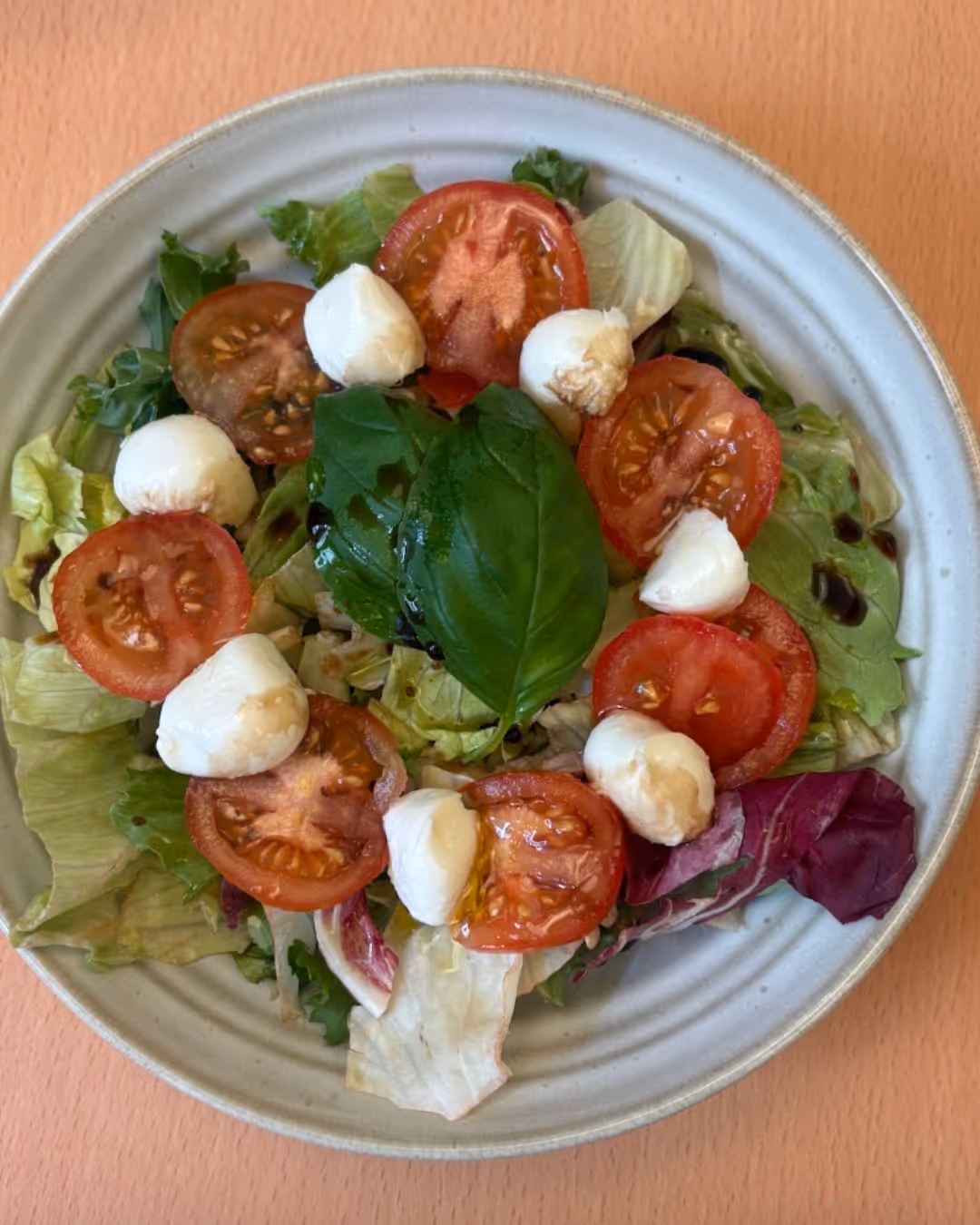 Fresh salad with mixed greens, tomato slices, small mozzarella balls, and a basil leaf on top, served on a white plate.