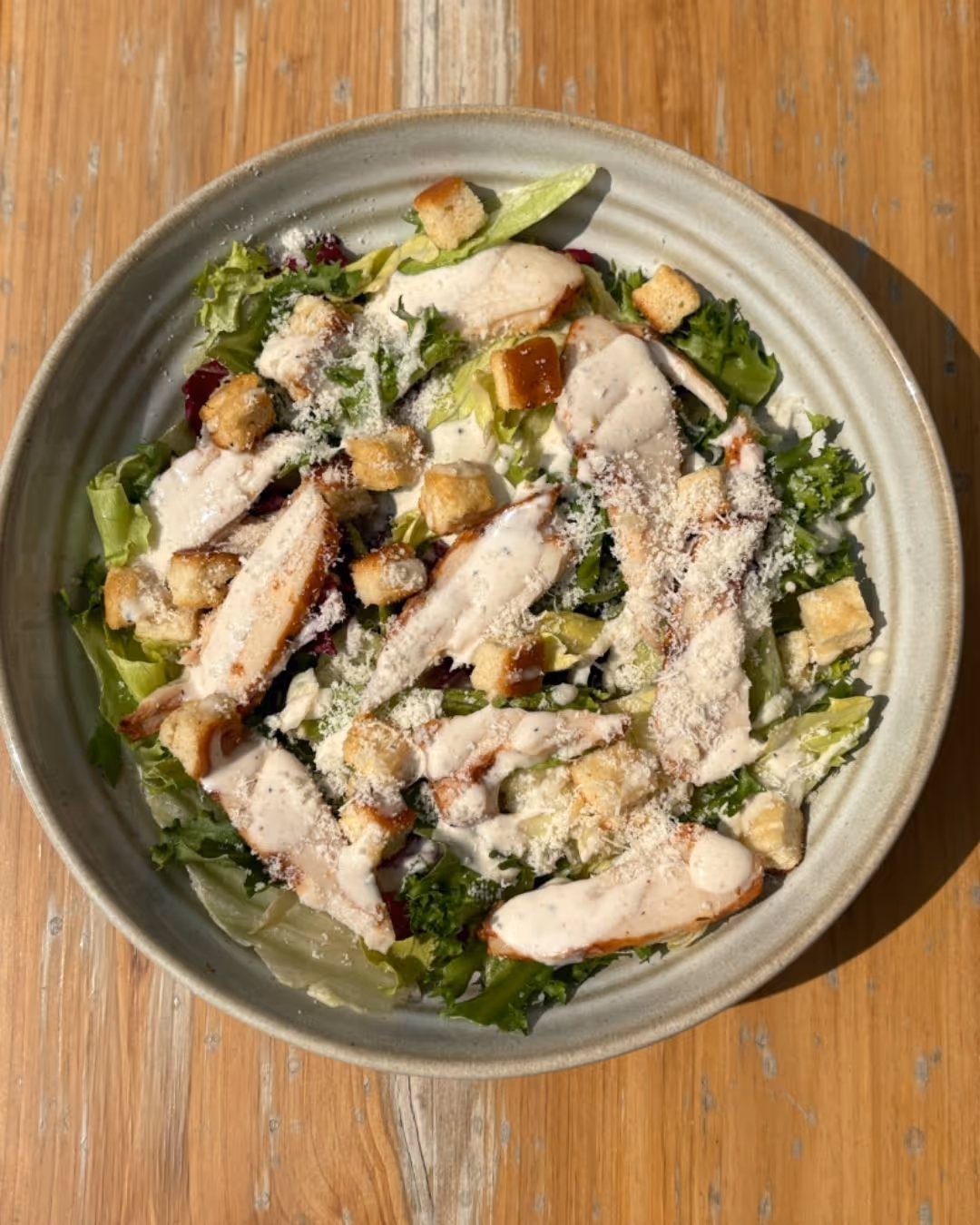 Caesar salad with grilled chicken strips, croutons, grated cheese, and creamy dressing on mixed greens in a ceramic bowl.