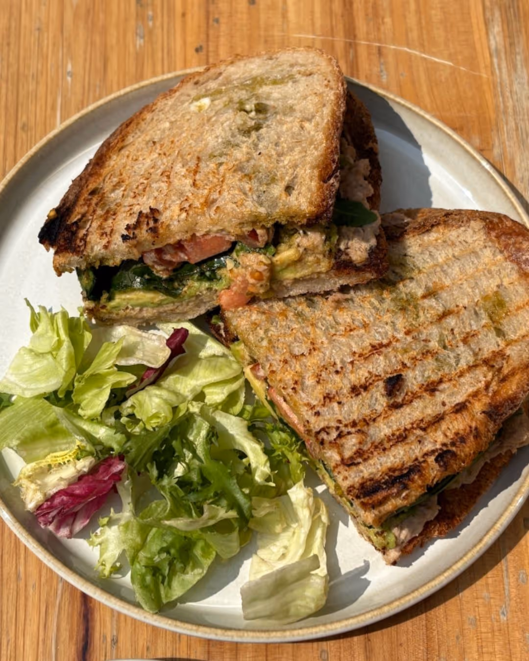 Grilled tuna melt sandwich cut in half with leafy green salad on a white plate on a wooden table.