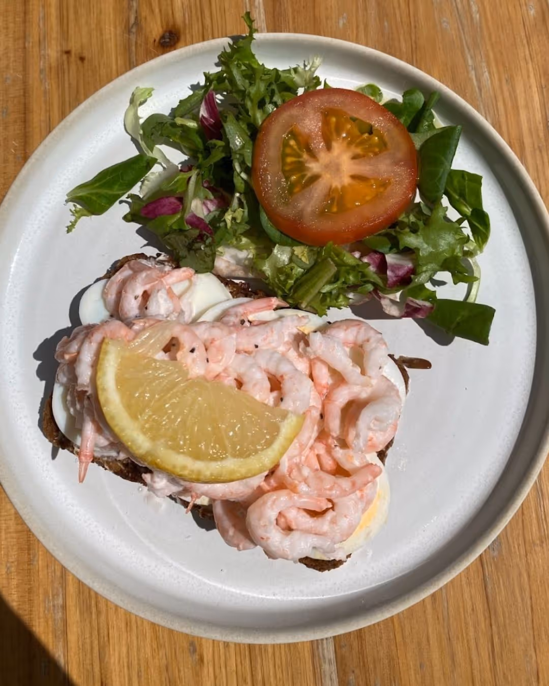 Open-faced shrimp sandwich topped with a lemon wedge, served with mixed greens and a slice of tomato on a white plate.