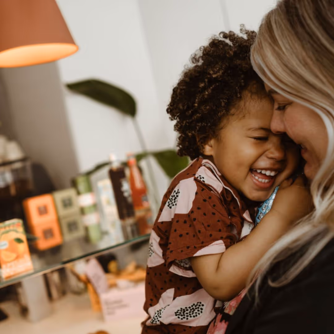 Young child with curly hair laughing joyfully while being hugged by an adult woman with blonde hair.