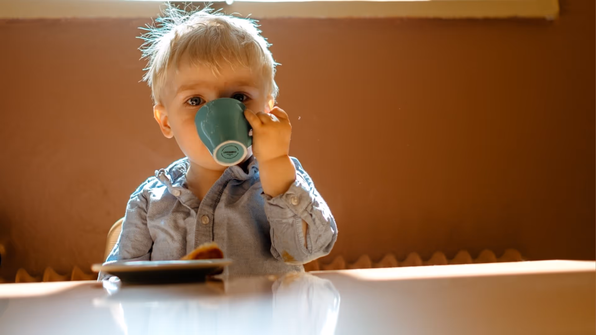 Young child with blonde hair drinking from a green cup at a table with a plate of food.