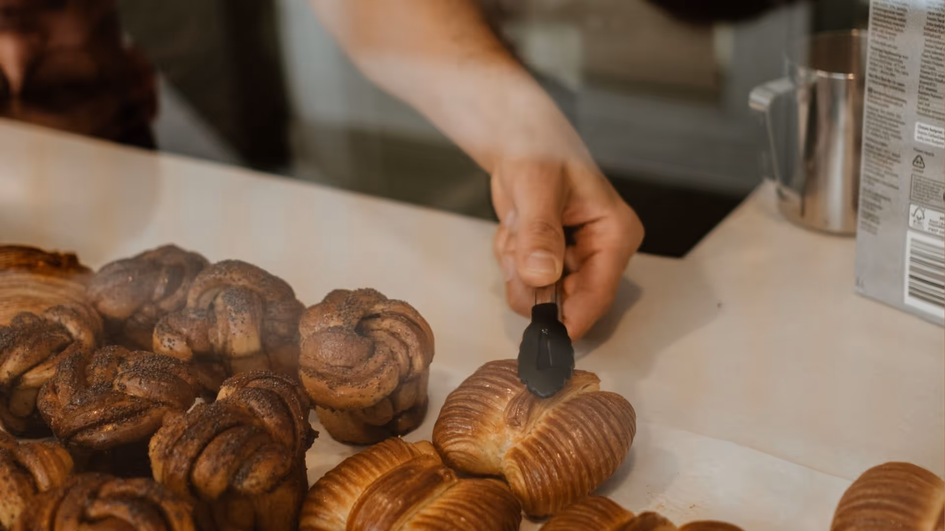Hand using black tongs to pick up a pastry among baked goods on a white countertop.
