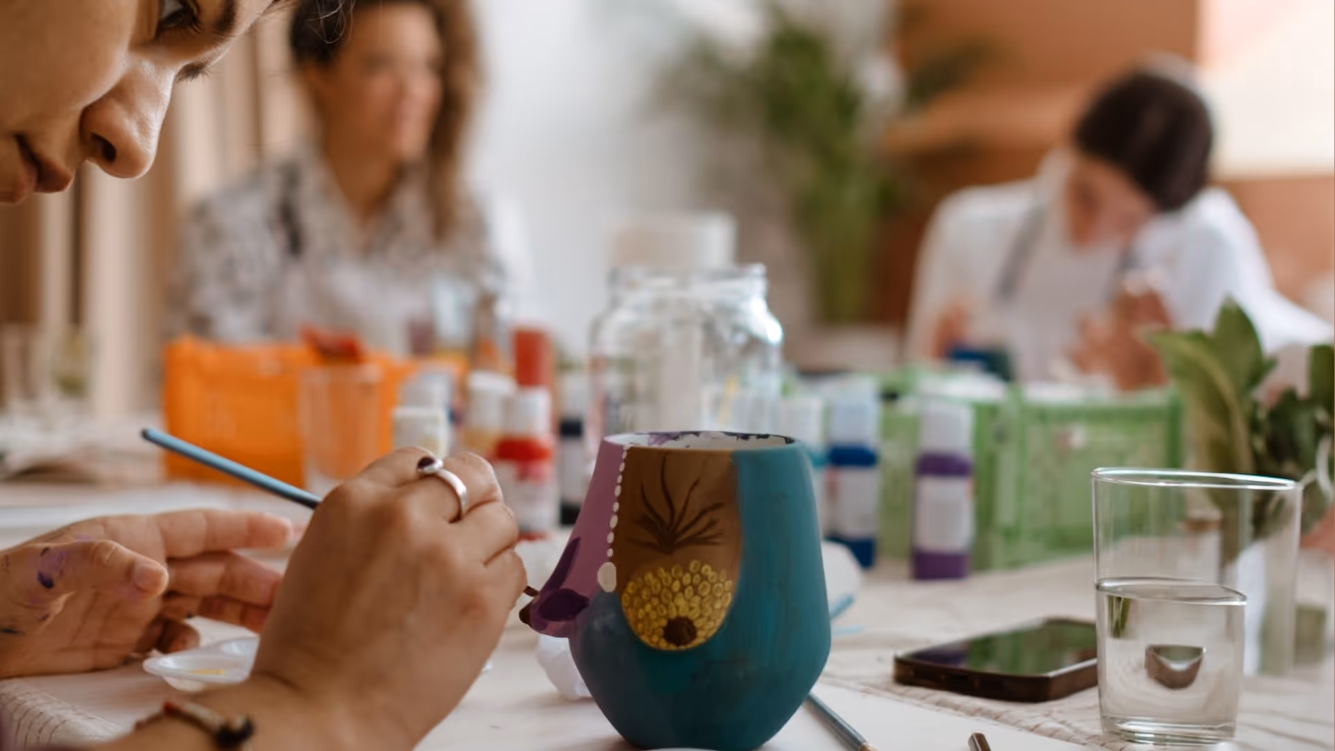 Person painting a colorful ceramic mug with art supplies on a table and blurred people in the background.