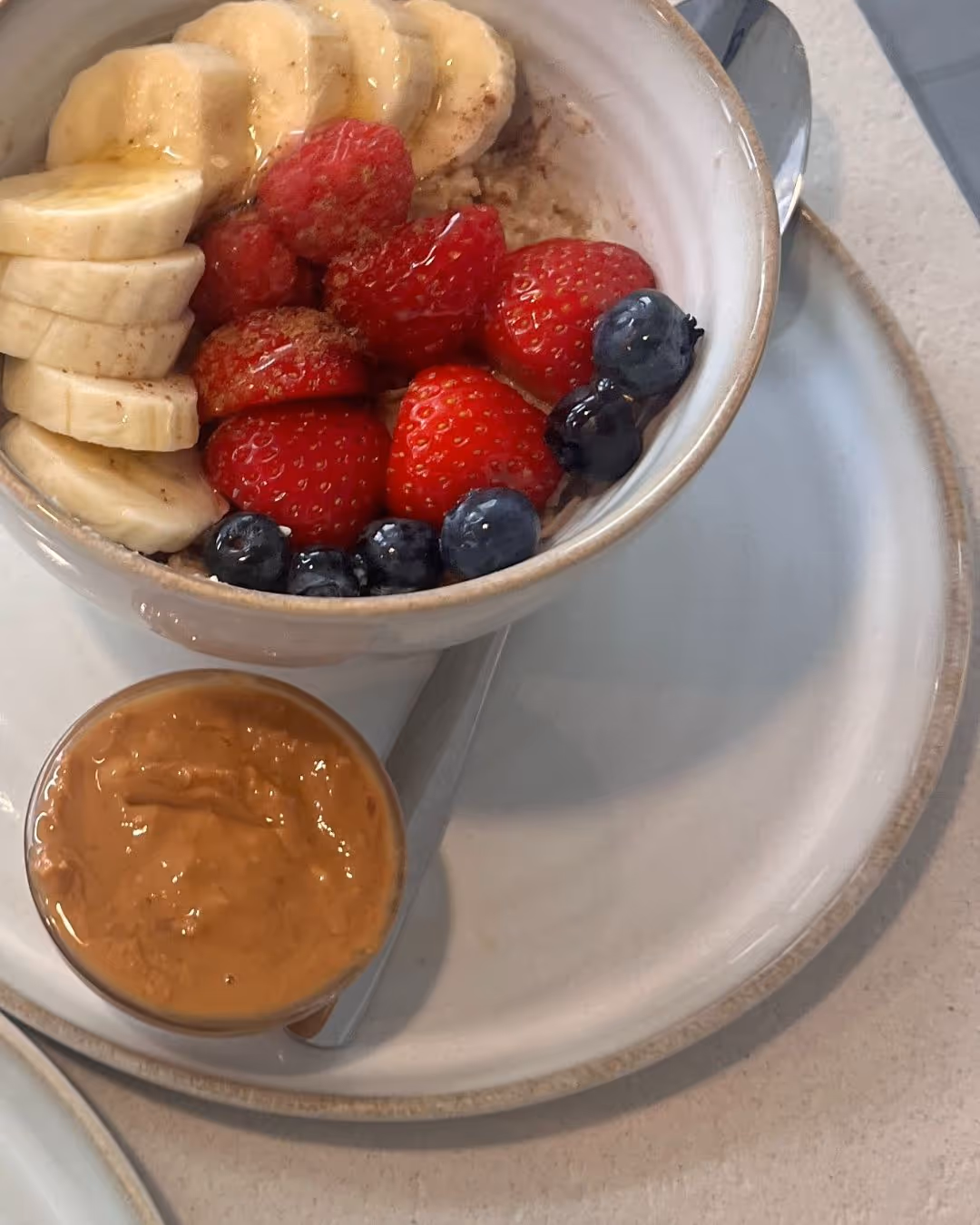 Bowl of oatmeal topped with sliced banana, strawberries, raspberries, blueberries, and a small cup of peanut butter on a beige plate with a spoon.