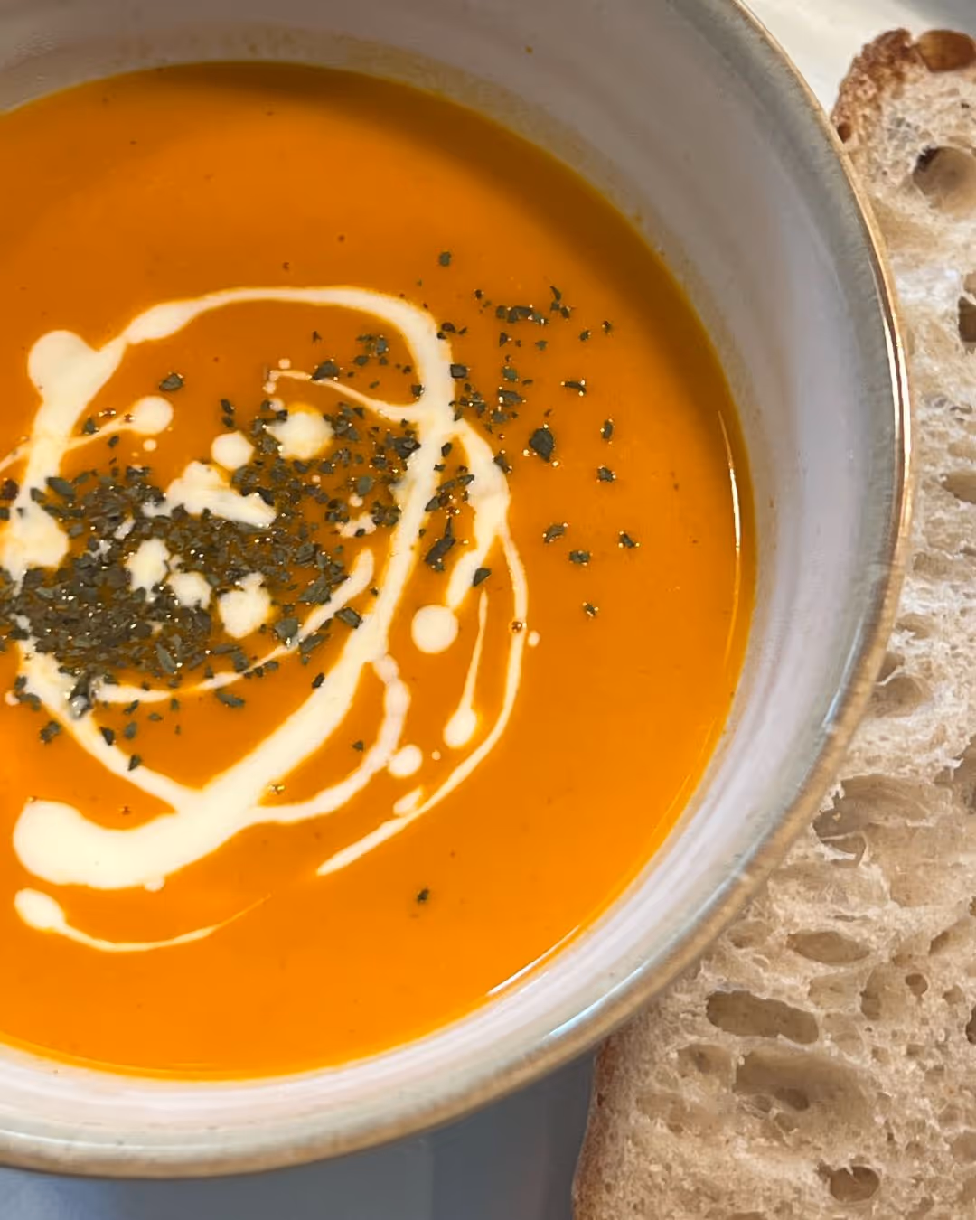 Bowl of creamy orange soup garnished with a swirl of cream and herbs, served with a slice of rustic bread.