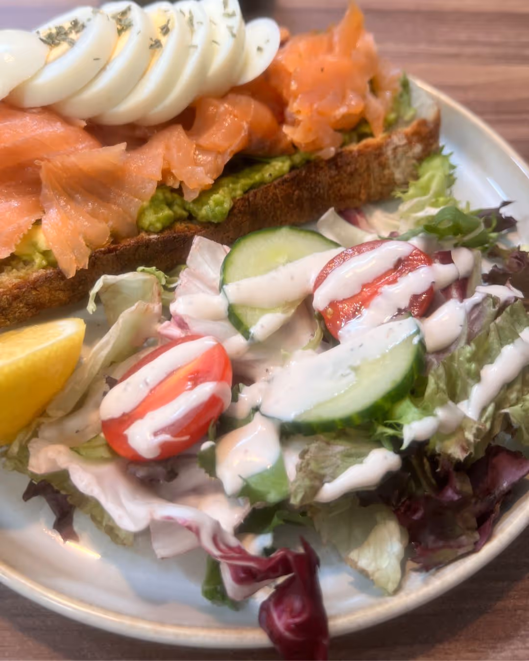 Plate with avocado toast topped with smoked salmon and sliced boiled egg, served with salad of lettuce, cherry tomatoes, cucumber slices, drizzled with dressing and a lemon wedge.