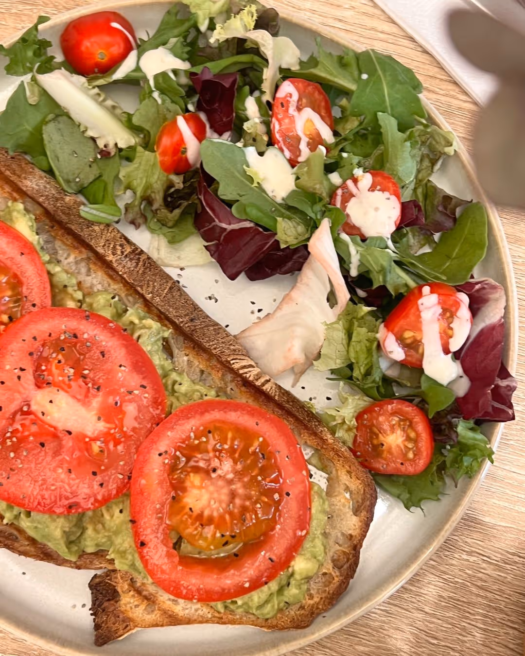 Plate with avocado toast topped with sliced tomatoes and black pepper, served with a side salad of mixed greens and cherry tomatoes drizzled with dressing.