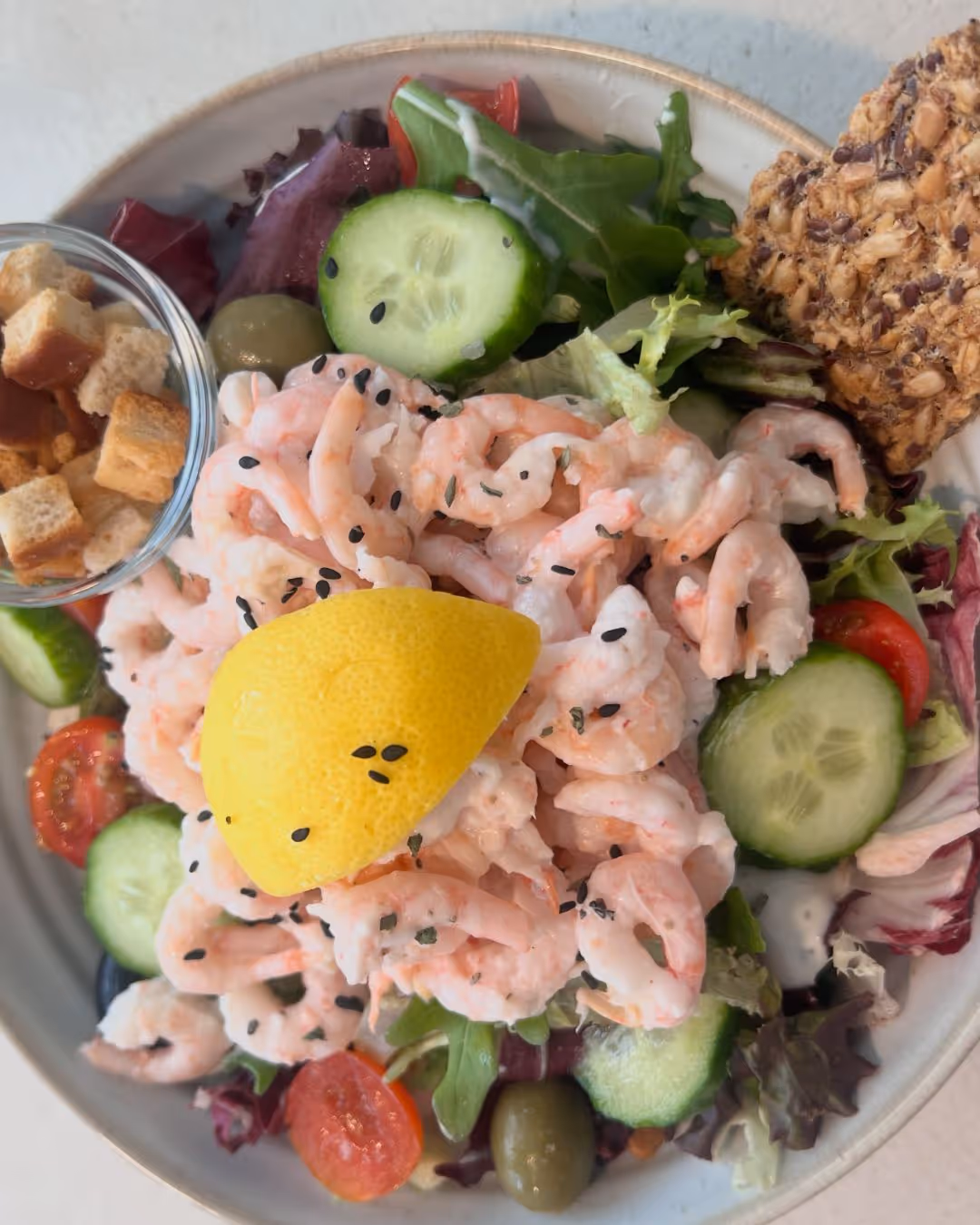 Prawn salad with mixed greens, cucumber slices, cherry tomatoes, green olives, a lemon wedge, black sesame seeds, croutons, and a seeded cracker.