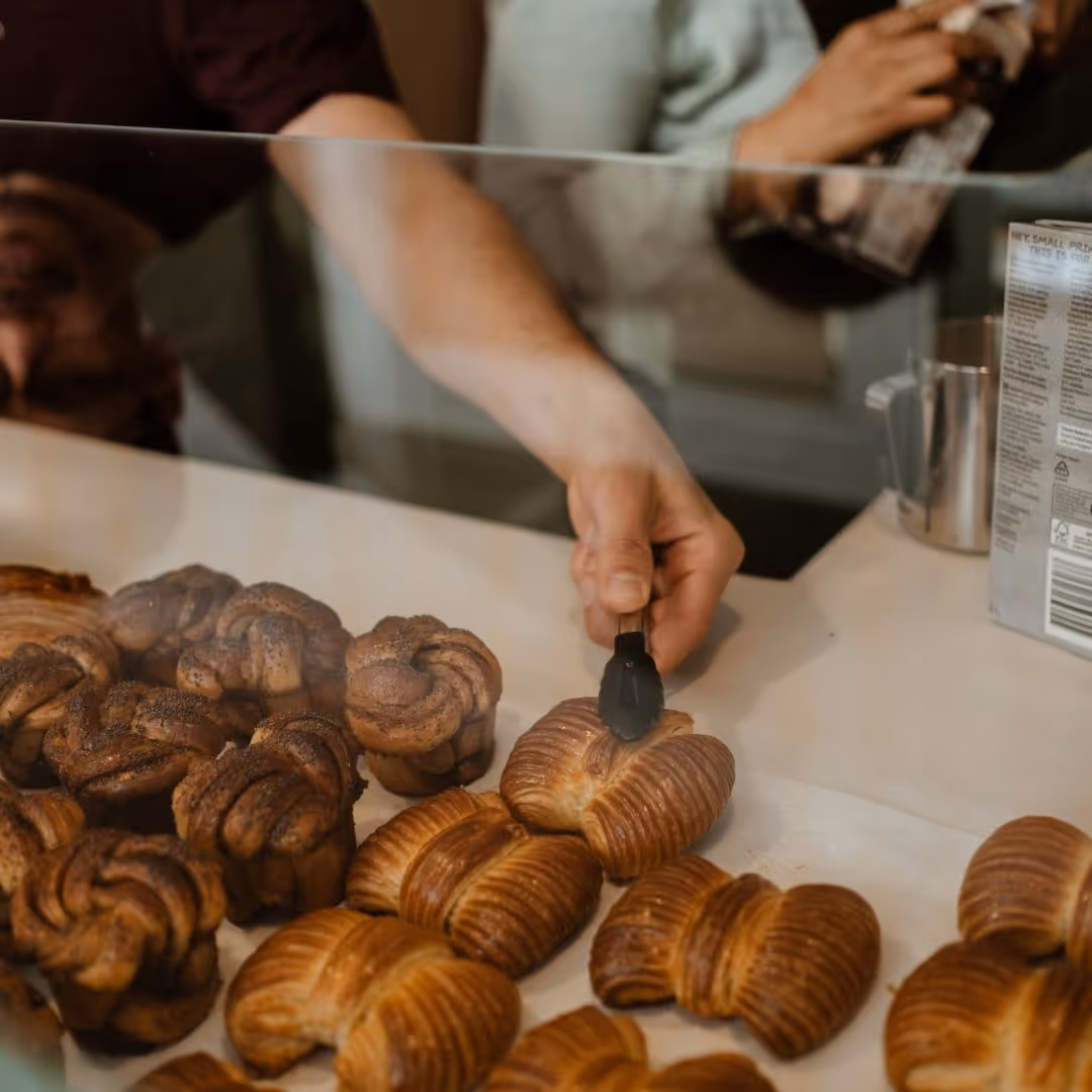 A man picking freshly baked rosette buns with tongs from a glass display counter, with a barista working in the background in café in Stockholm