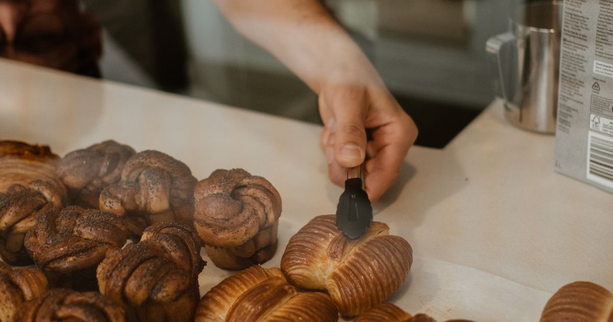 A man picking freshly baked rosette buns with tongs from a glass display counter, with a barista working in the background in café in Stockholm