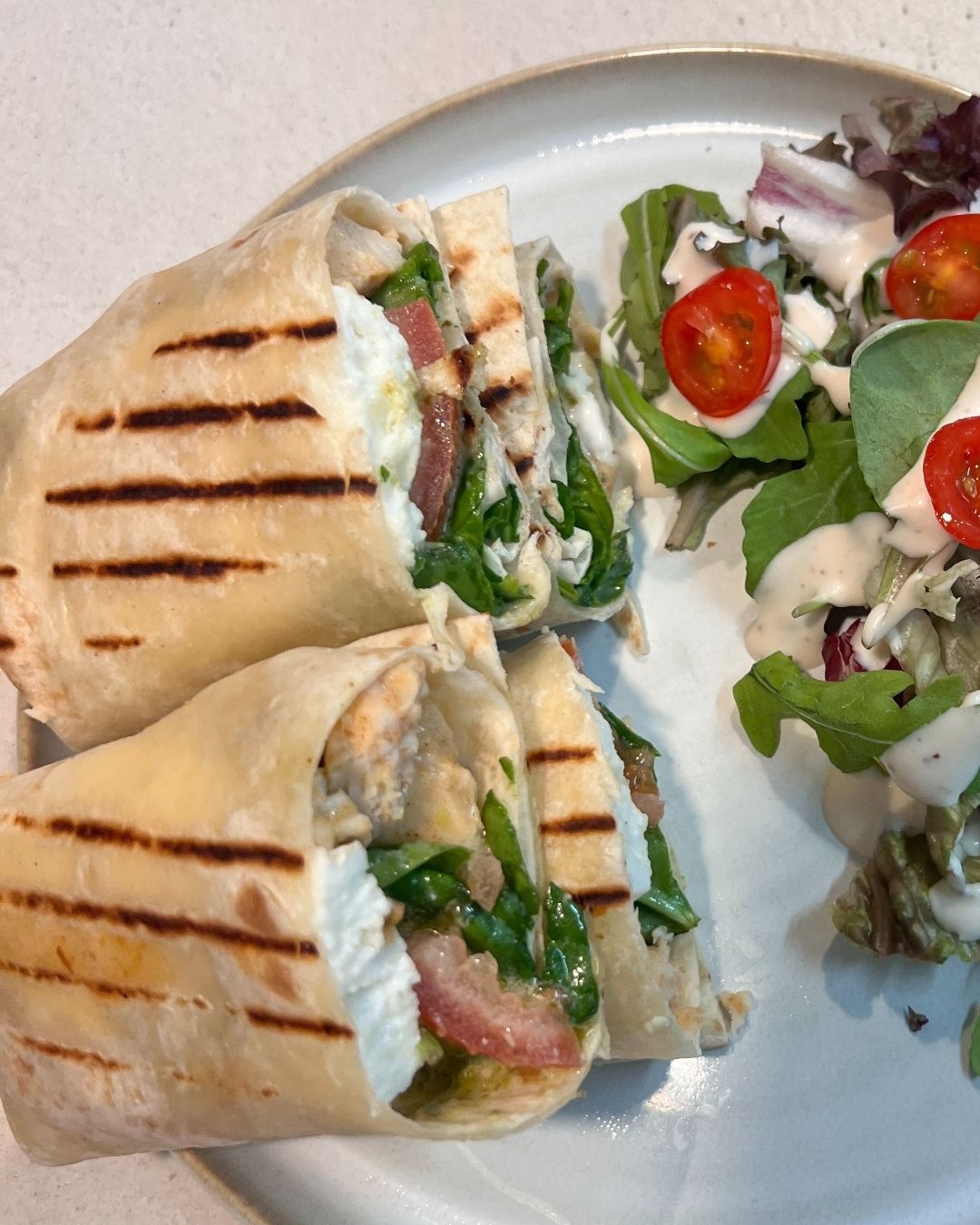 Grilled wrap cut in halves filled with spinach, tomato, cheese, and other ingredients served with a side salad of leafy greens and cherry tomatoes with dressing.