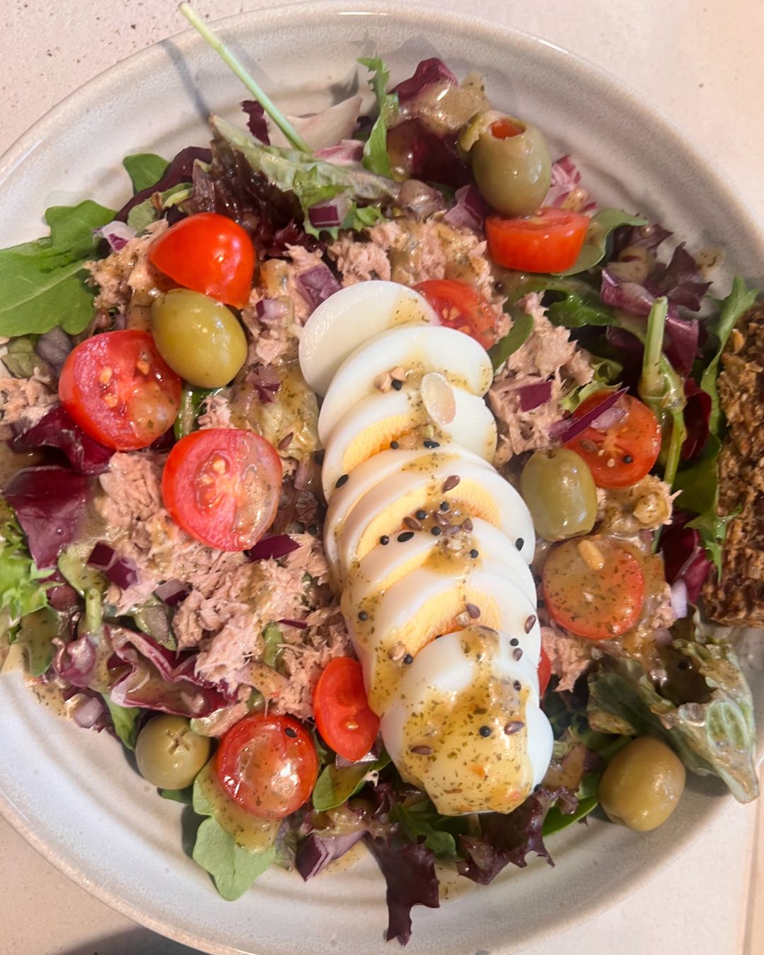 Plate of tuna salad with mixed greens, halved cherry tomatoes, green olives, sliced boiled egg, and sprinkled seeds with dressing.