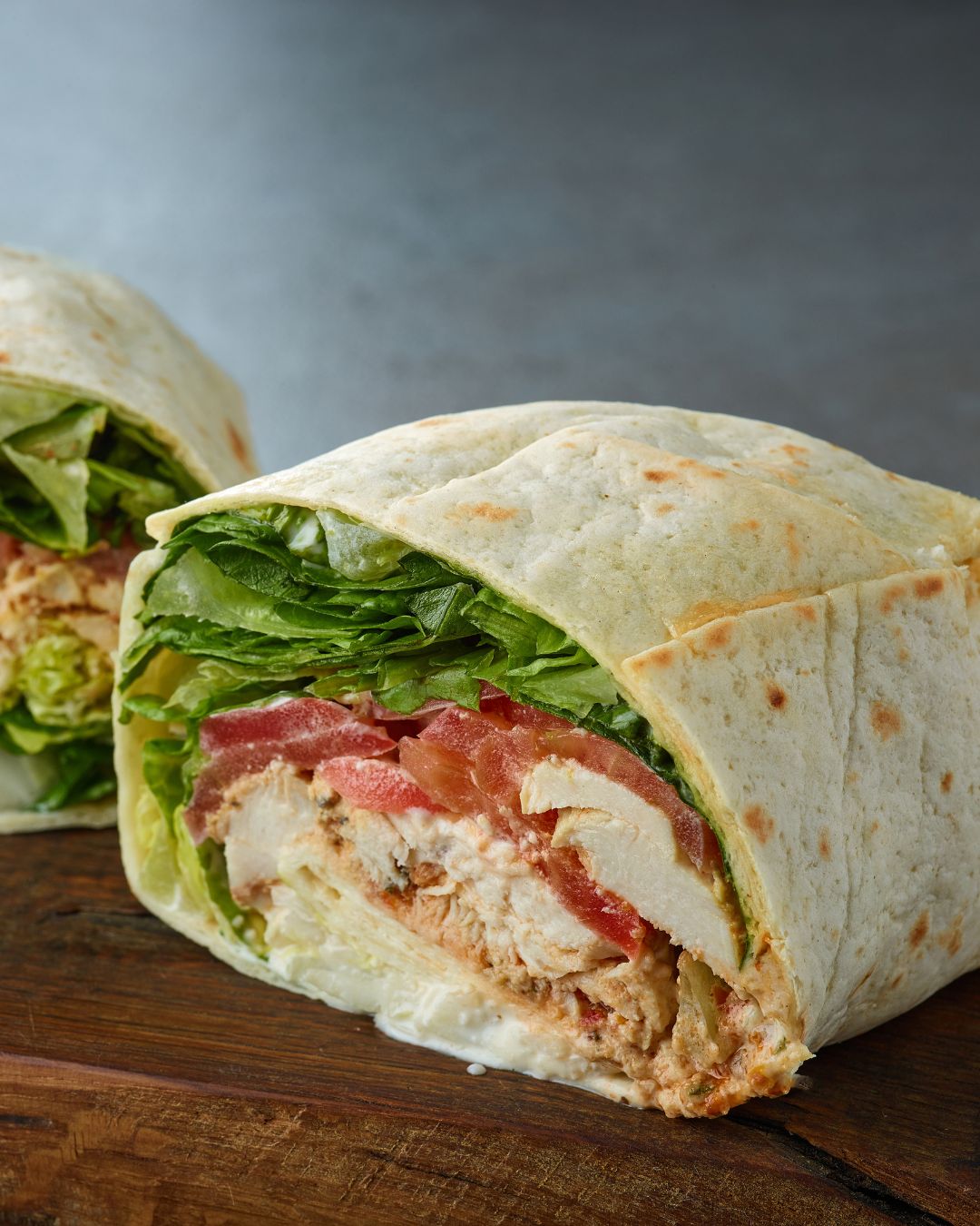 Close-up of a chicken wrap cut in half showing grilled chicken, lettuce, tomato, and mayonnaise in a flour tortilla on a wooden surface.