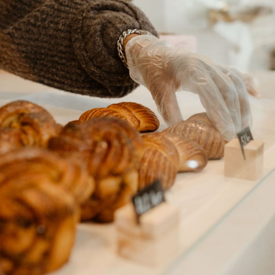 Gloved hand picking up a freshly baked, golden brown pastry from a display case filled with various baked goods.