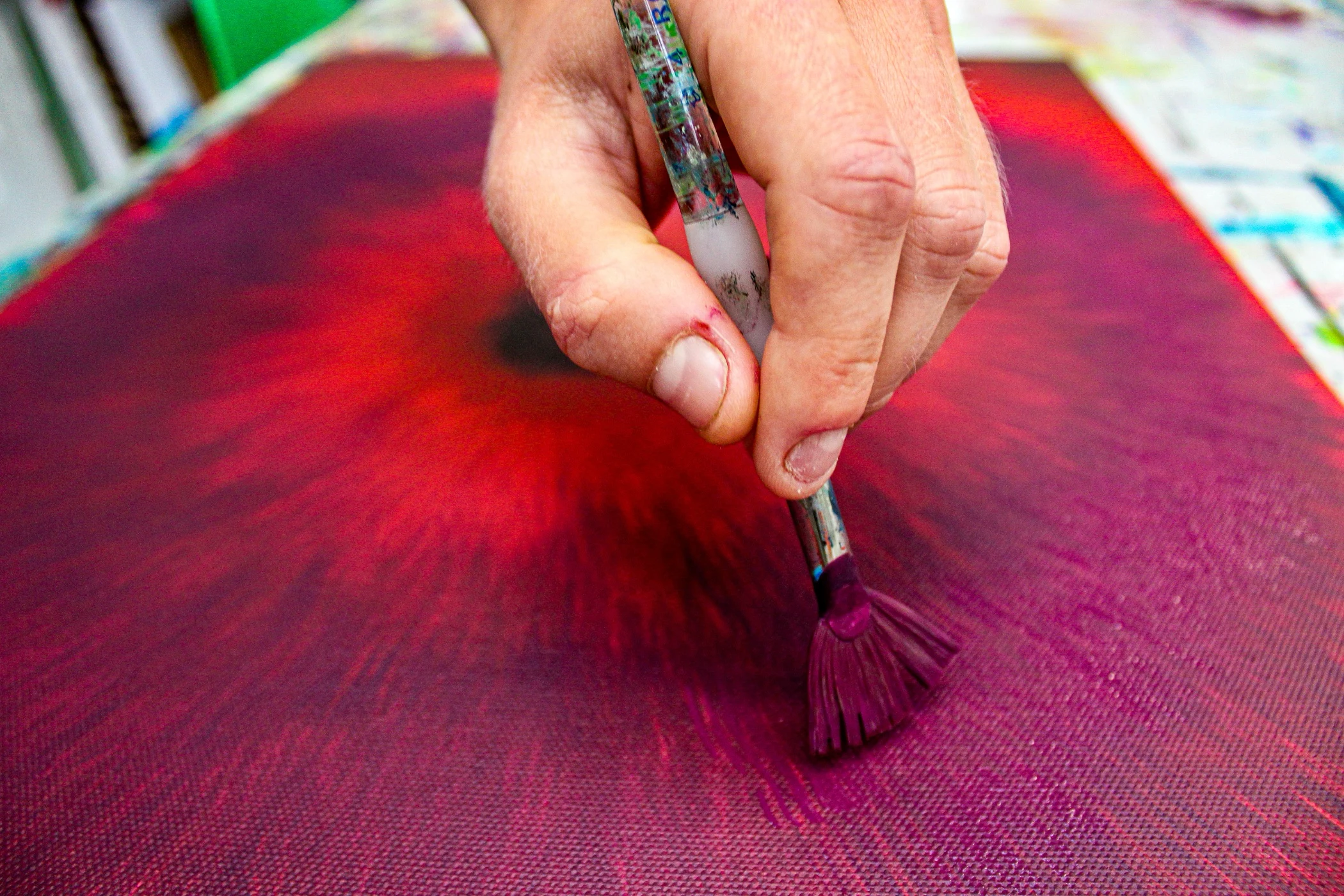 Close-up of a hand holding a paintbrush applying purple paint on a textured canvas with red and purple hues.