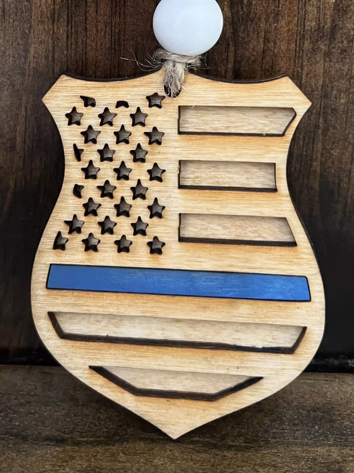 Wooden badge shaped Thin Blue Line flag with carved stars and stripes hanging by a rope.