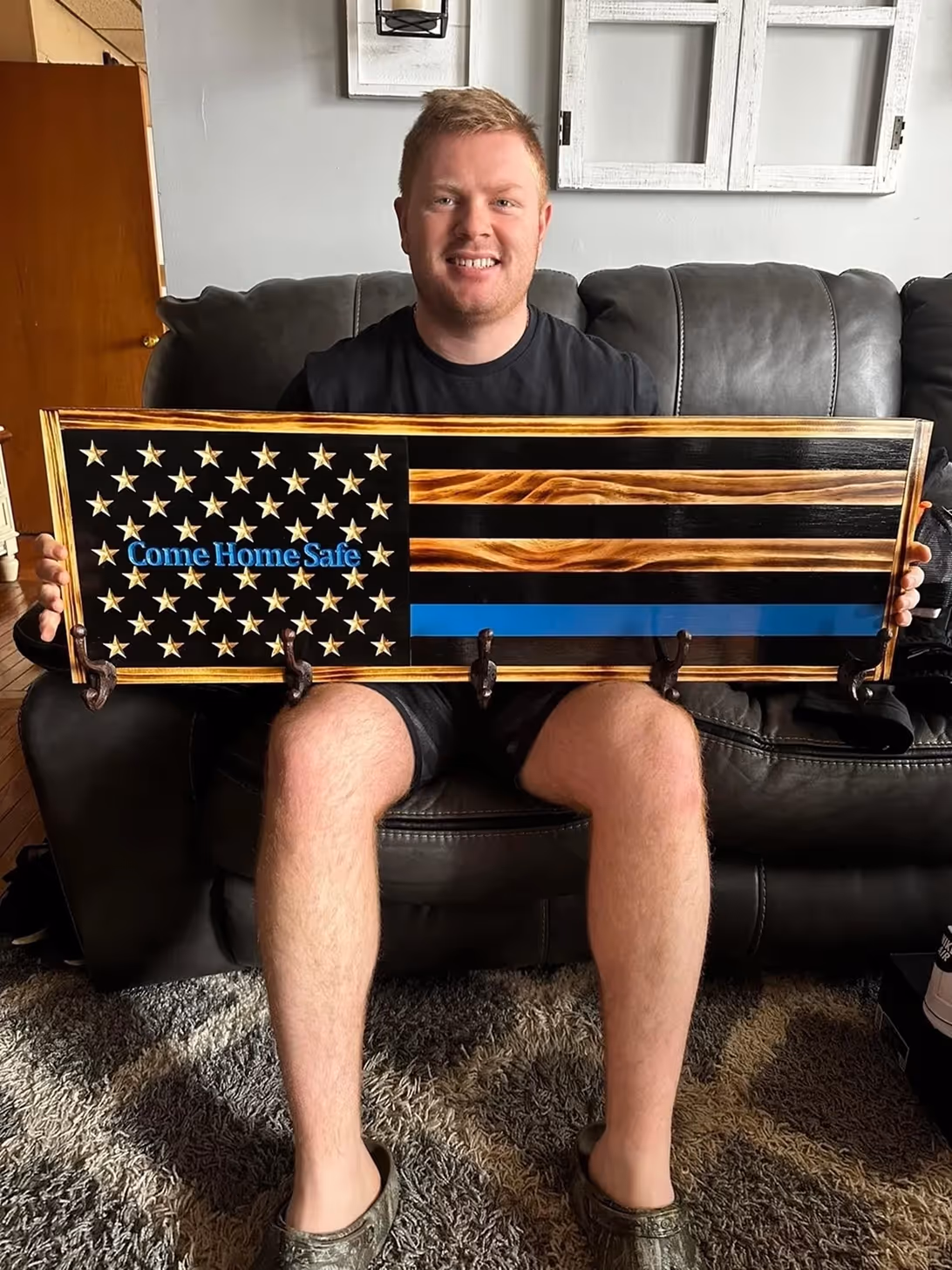 Man sitting on couch holding wooden American flag with 'Come Home Safe' and a thin blue line.