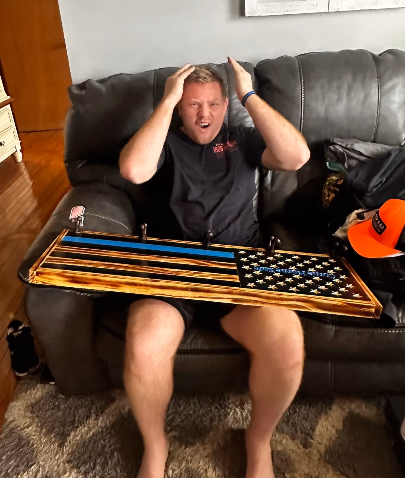 Man on couch holding wooden American flag plaque with thin blue line and hooks.