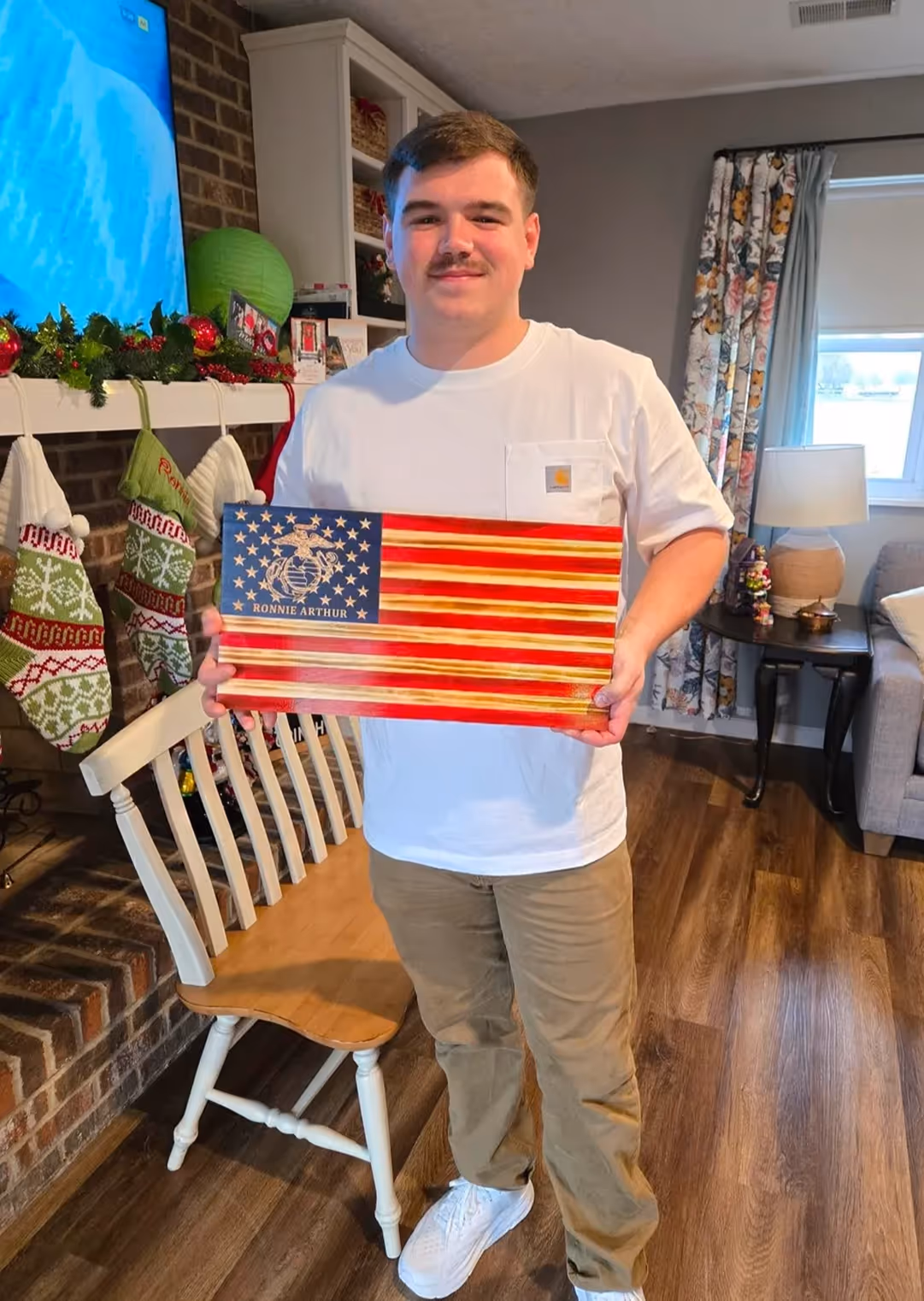 Young man holding American flag plaque with Marine Corps emblem and name Ronnie Arthur in living room.