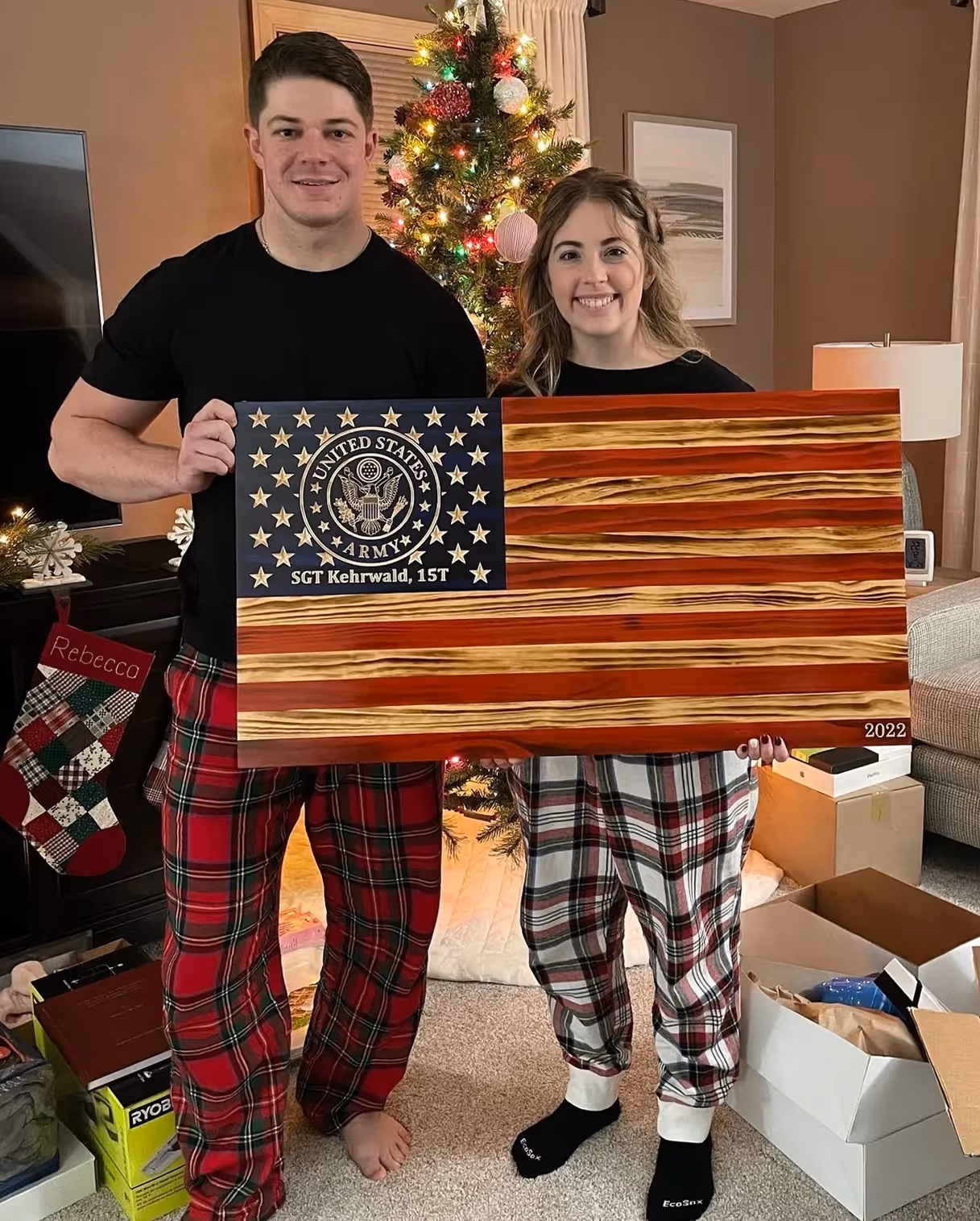 Man and woman in plaid pajama pants holding wooden US Army flag plaque by Christmas tree.
