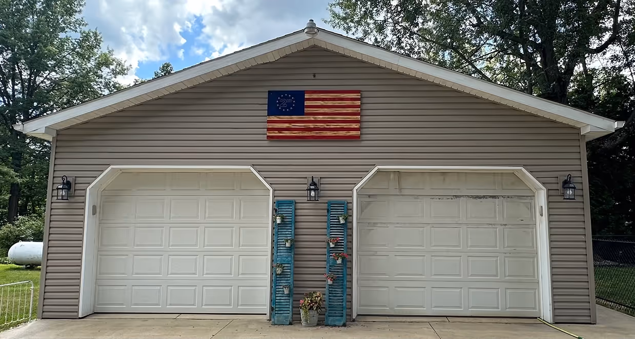 Two white garage doors on a beige building with a 2nd Amendment flag and blue shutters holding plants.