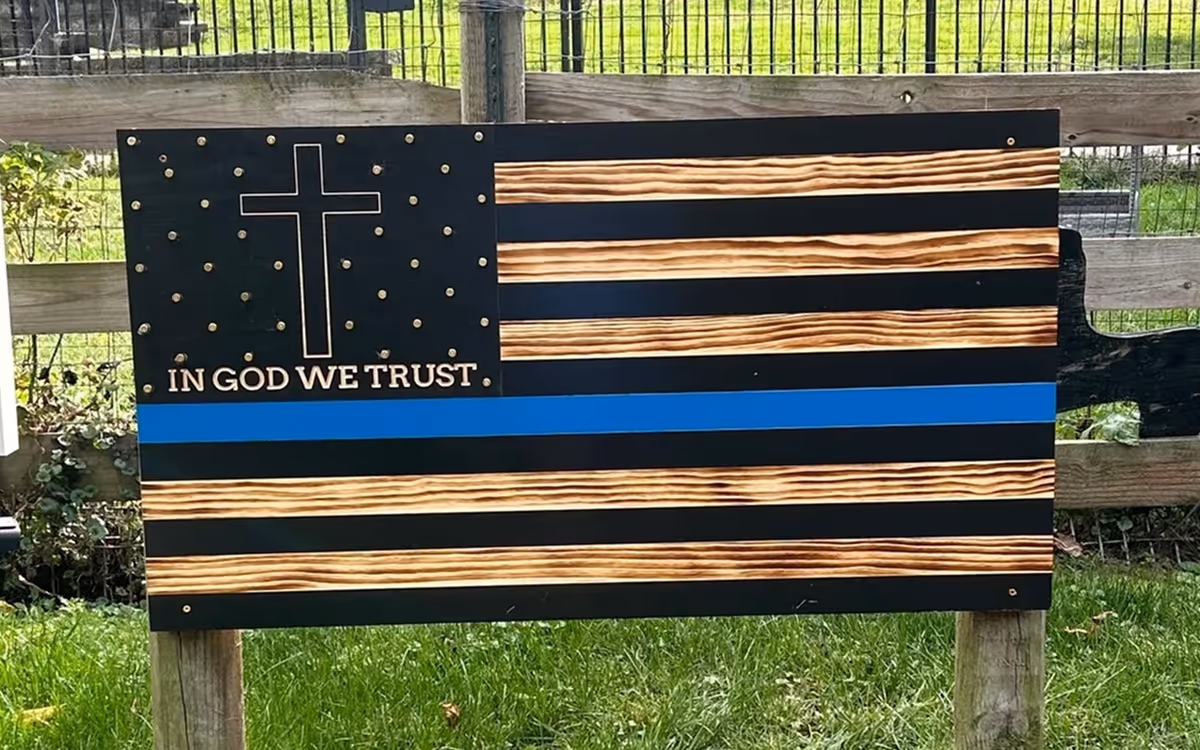 Wooden American flag with a blue stripe, a cross, and text In God We Trust on stars section.
