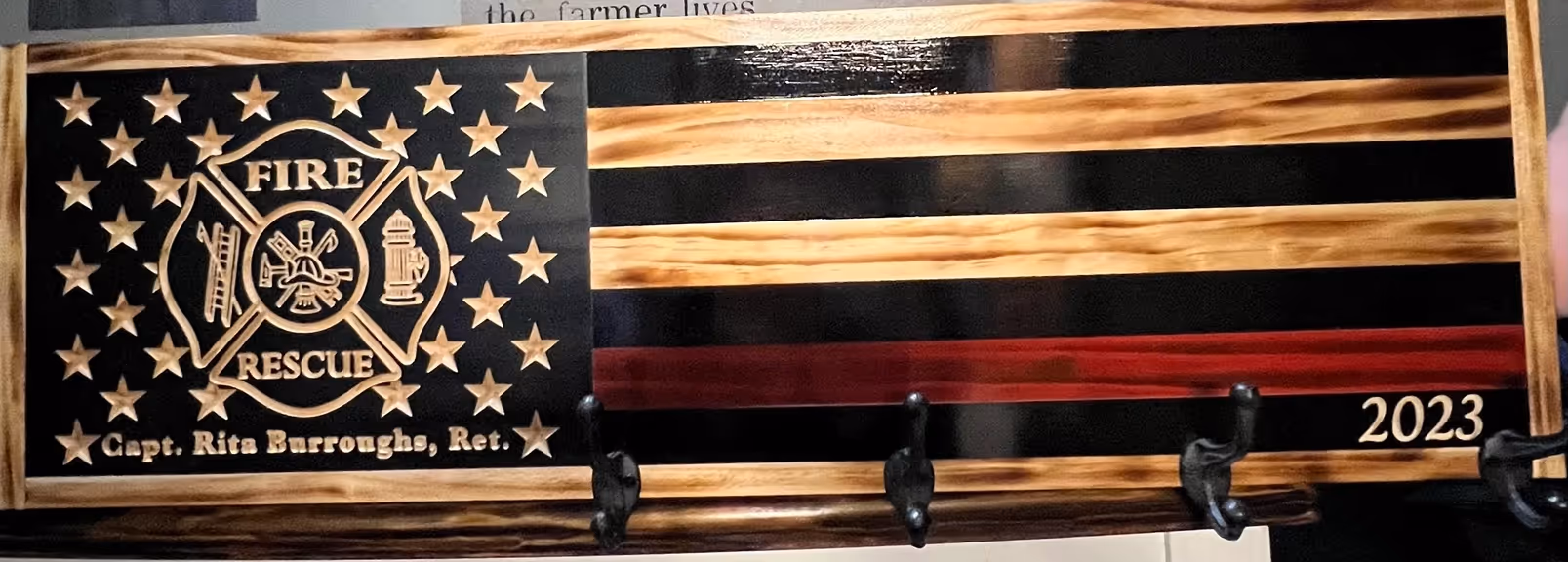 Wood flag plaque with fire rescue emblem, name Capt. Rita Burroughs Ret., and year 2023 with hooks below.