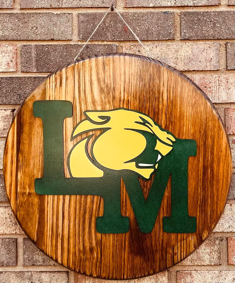 Round wooden sign with green letters LM and a yellow panther head between them, hanging on a brick wall.