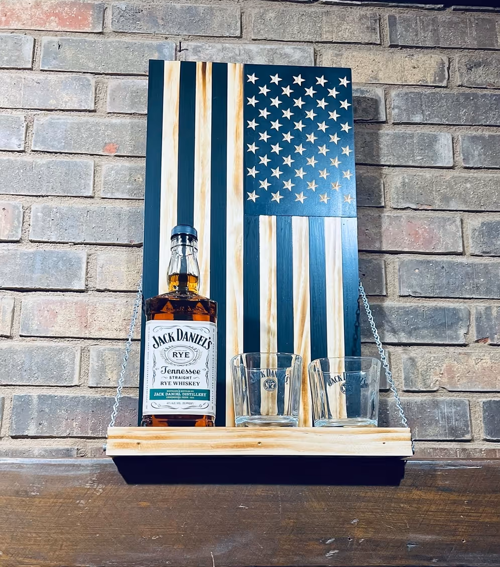 Jack Daniel's Tennessee Rye whiskey bottle and two branded glasses on a wooden shelf with an American flag backdrop.