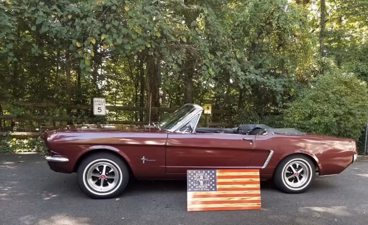 Maroon vintage convertible car parked on pavement near a wooden fence and green trees.